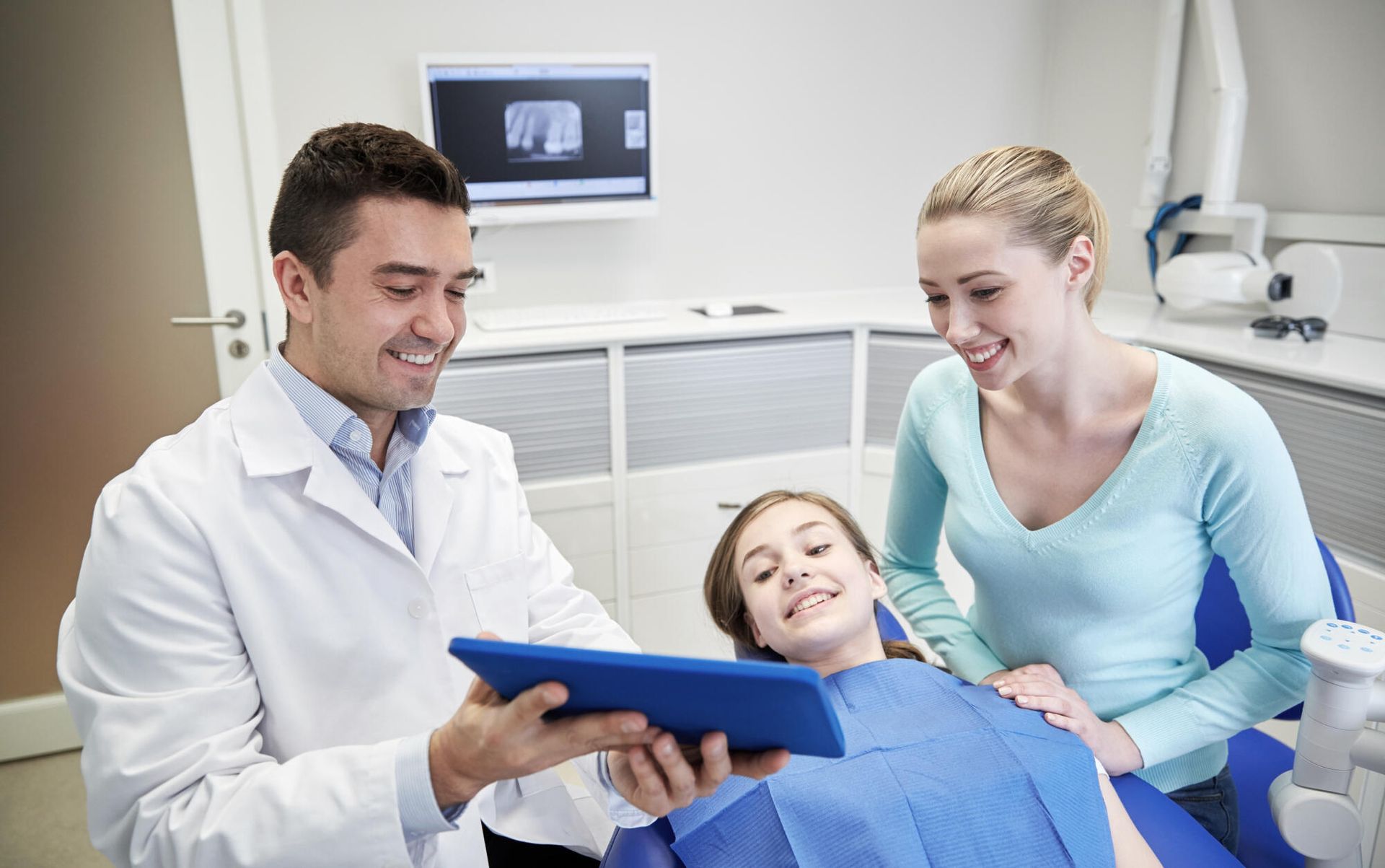 Dentist showing a blue tablet to a smiling patient while an assistant stands beside her in a dental clinic