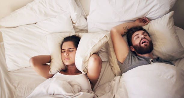 A man is snoring while a woman covers her ears in bed.