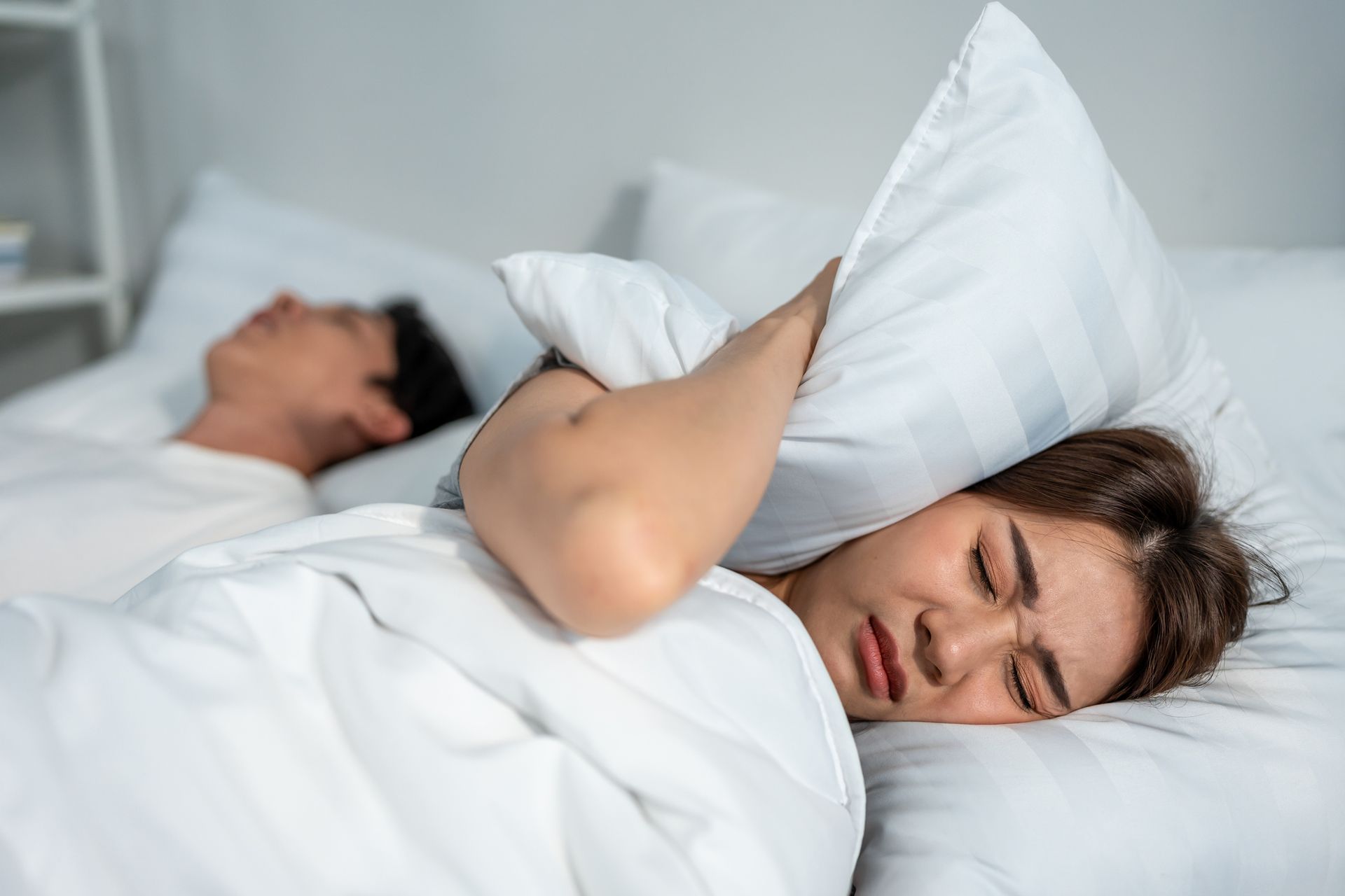 A woman is covering her ears with a pillow while a man sleeps next to her. A woman is covering her ears with a pillow while a man sleeps next to her.