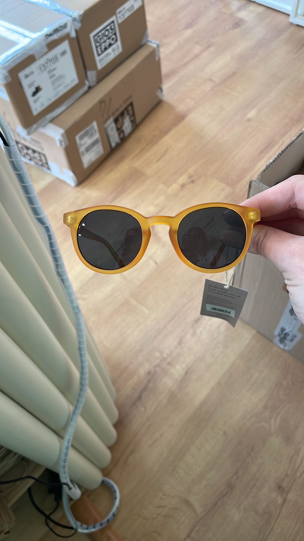 Una mano tiene in mano un paio di occhiali da sole rotondi con montatura gialla, in casa, su un pavimento di legno vicino a delle scatole