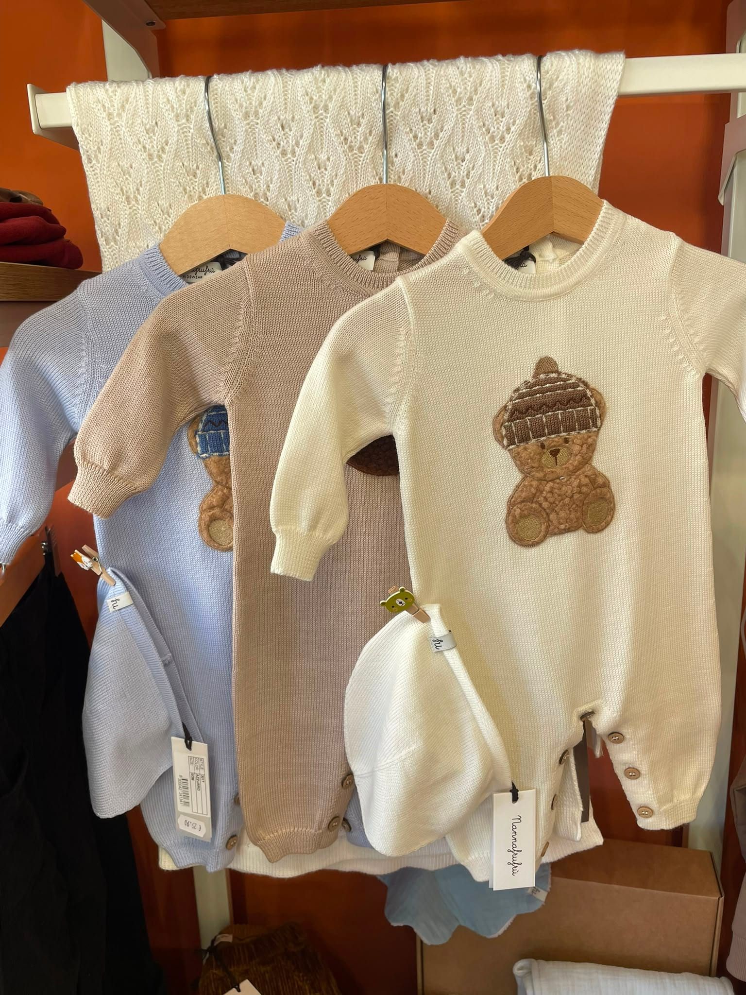 Tre tutine per neonati appese alle grucce, tra cui completini in maglia color crema e blu, esposte su un appendiabiti di legno.