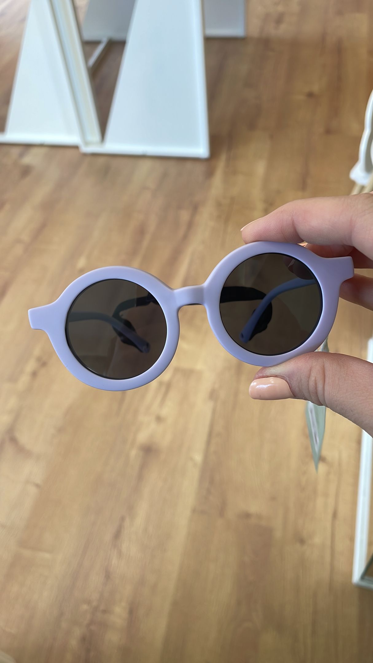 Mano che tiene un paio di occhiali da sole rotondi color lavanda sopra un pavimento di legno, in un ambiente interno