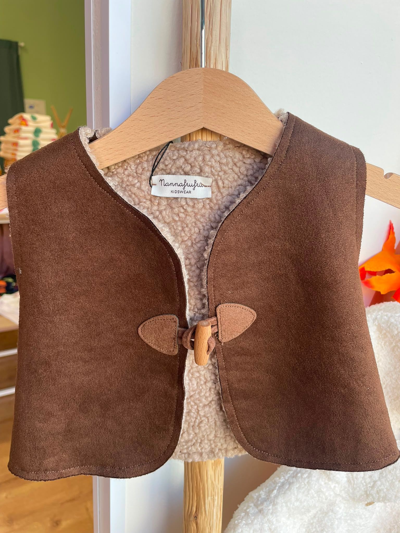 Gilet di lana marrone su una gruccia di legno, esposto in un ambiente interno su uno sfondo delicatamente illuminato.