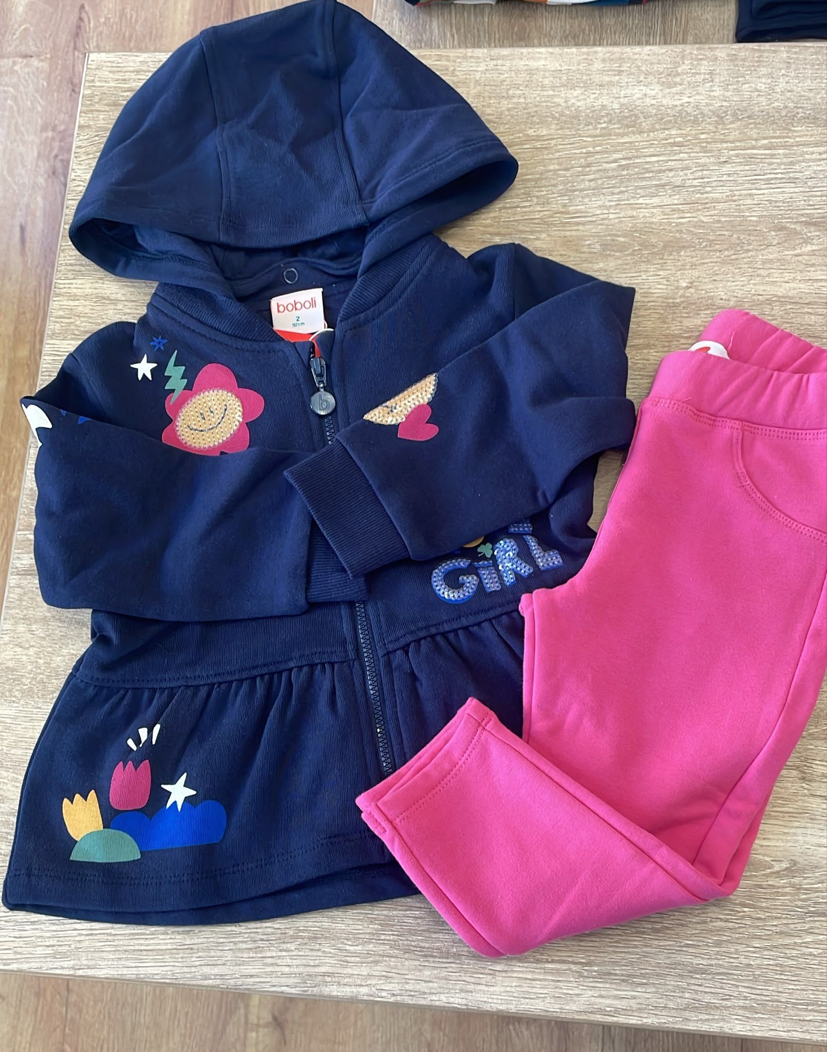 Completo blu con cappuccio per bimbo, con ricami floreali, accanto a pantaloni rosa su un tappeto