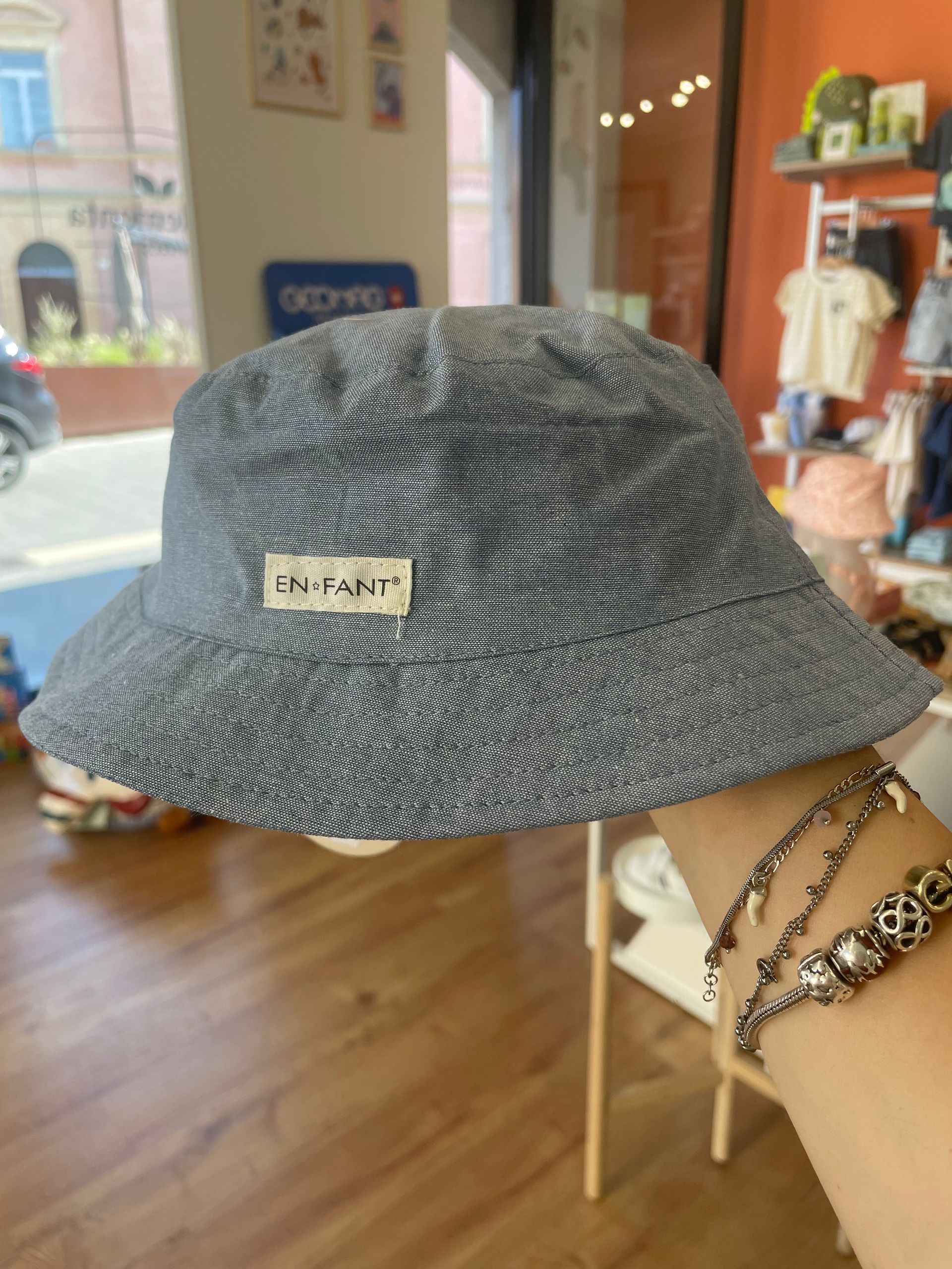 Una mano tiene en alto un sombrero de pescador gris estampado con una pequeña etiqueta, en una habitación con suelo y estanterías de madera.