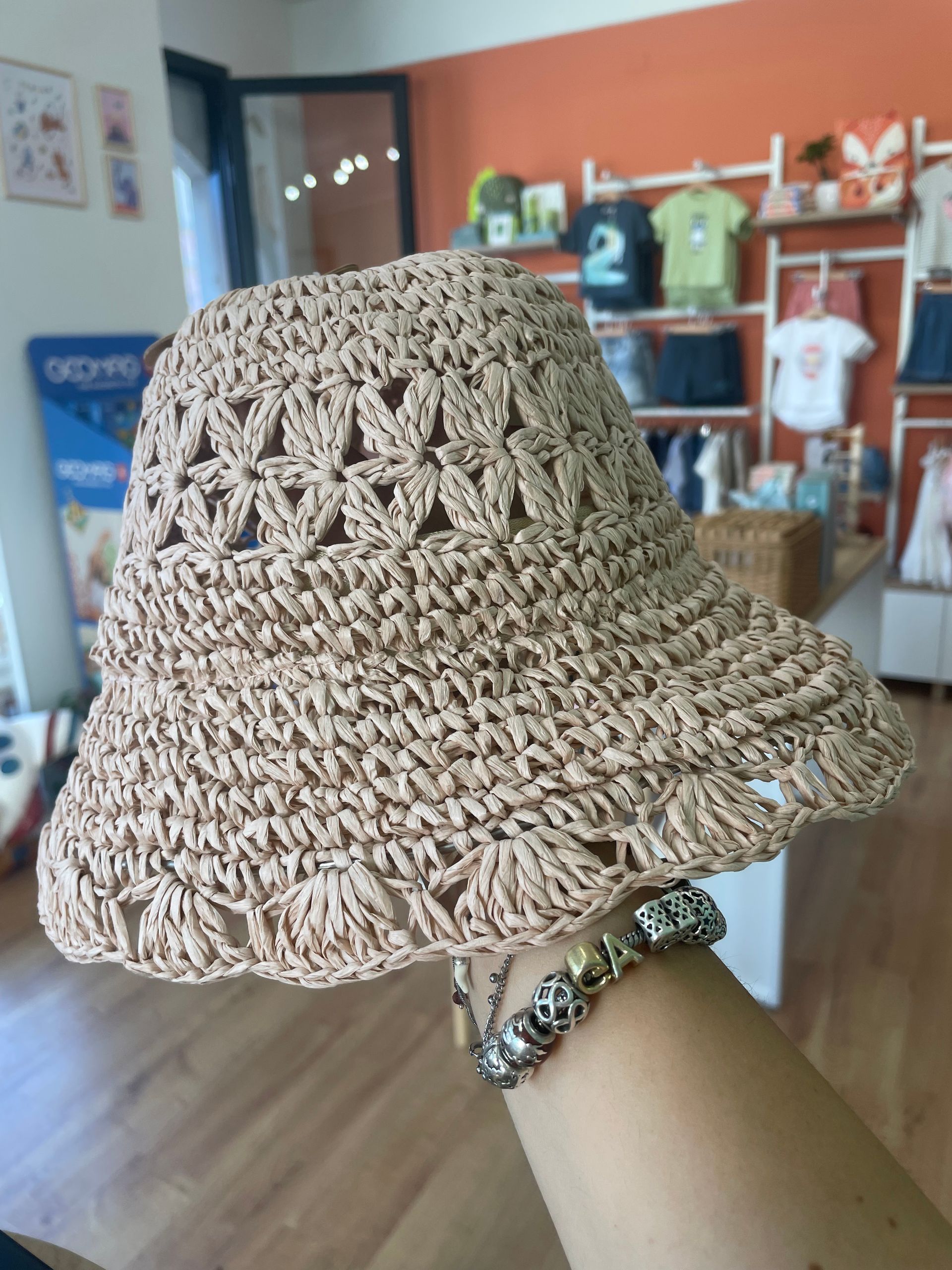Cappello parasole beige lavorato all’uncinetto a mano, tenuto in alto al chiuso in una stanza con scaffali e una porta sullo sfondo