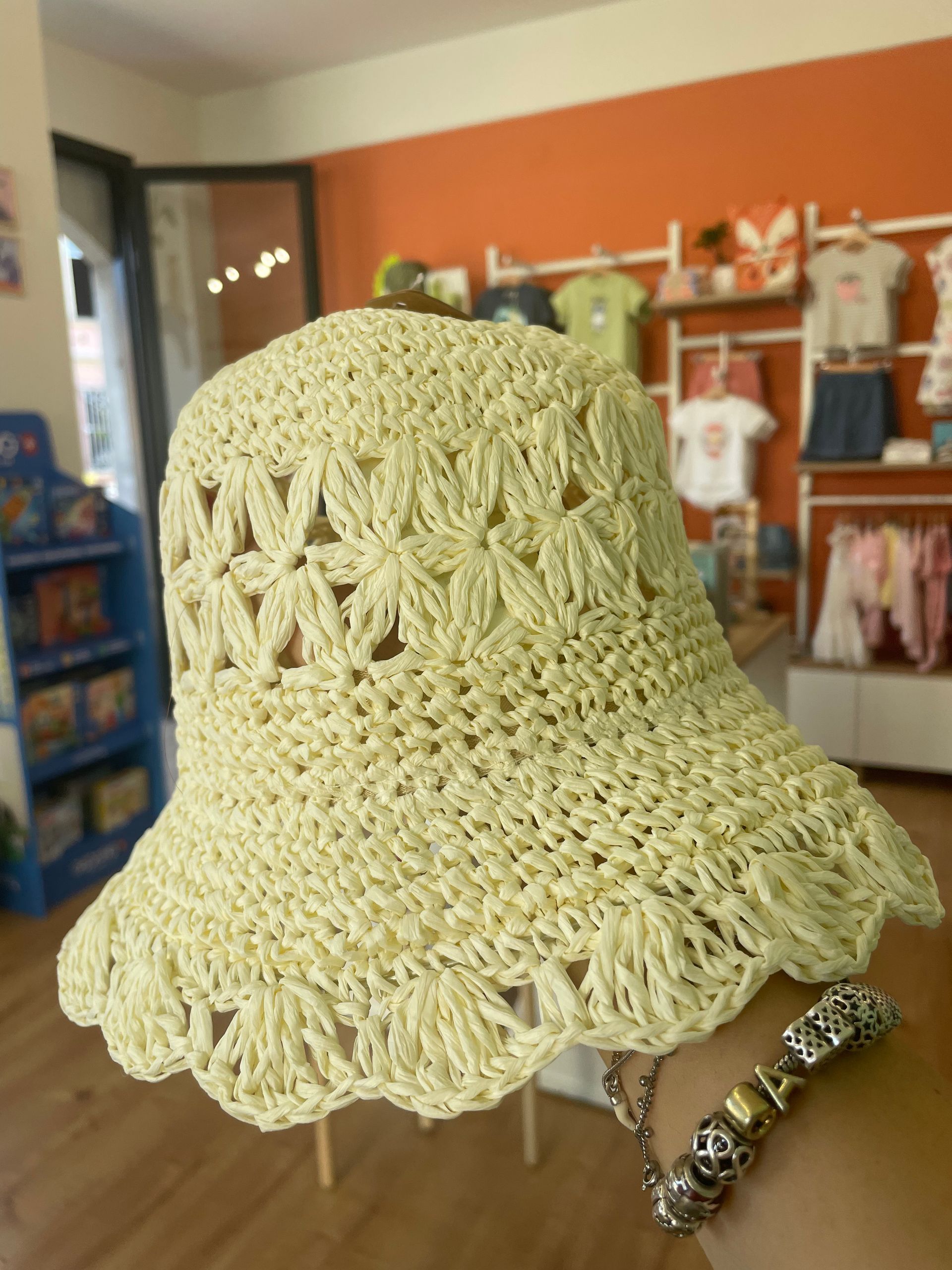 Primo piano di un cappello da sole color crema, lavorato all’uncinetto e con la tesa smerlata, tenuto in mano all’interno di un negozio di abbigliamento.
