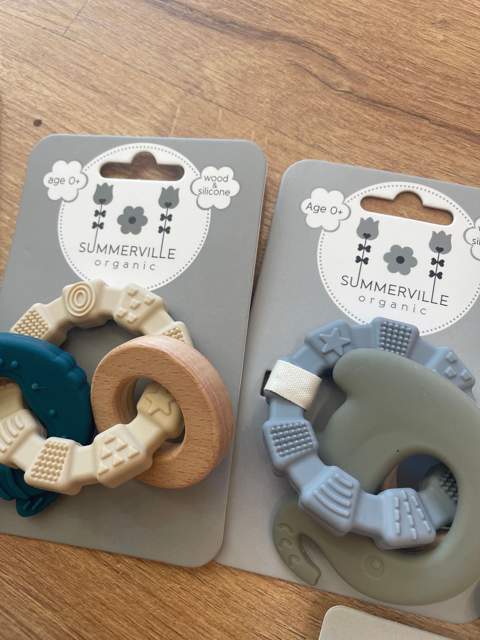 Due giocattoli da dentizione per neonati su cartoncini espositivi, uno beige e blu e l’altro grigio e blu, su una superficie di legno