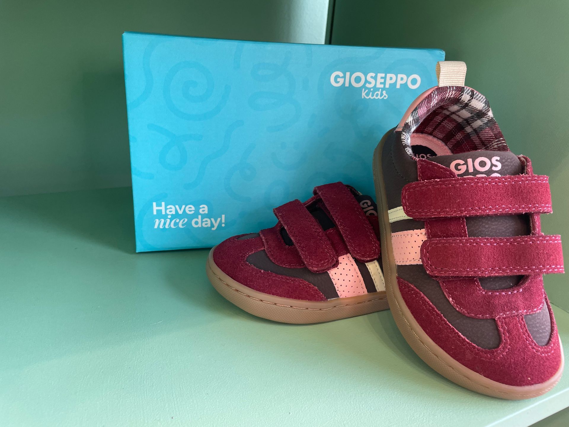 Scarpine rosse da bimbo con chiusure in velcro accanto a una scatola blu GIOSEPPO su sfondo verde