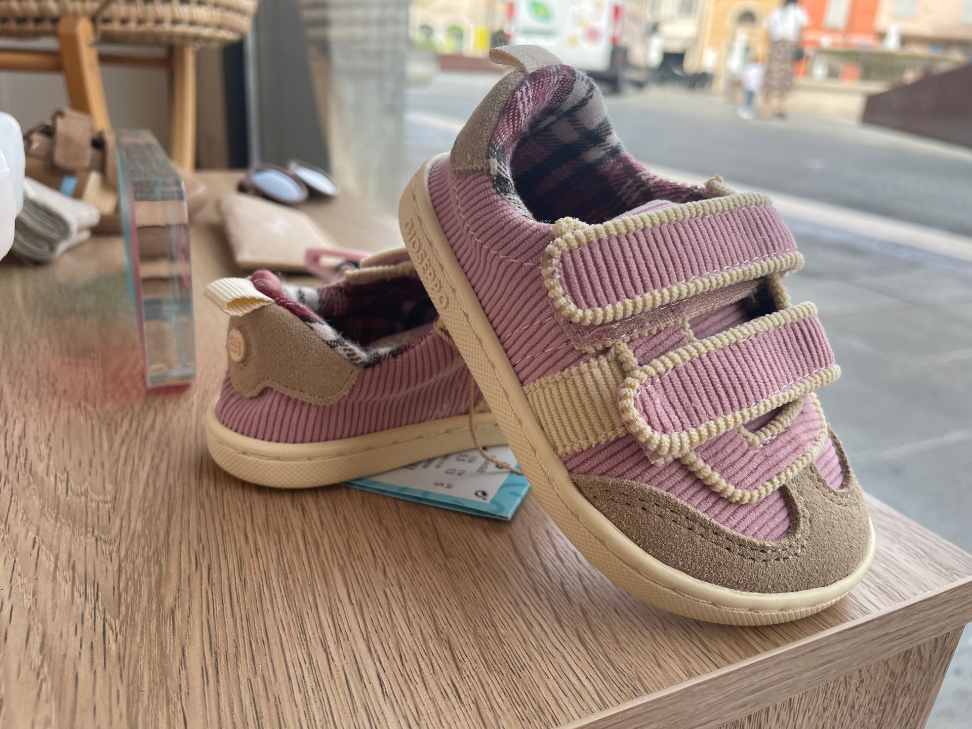 Sandali rosa da bimba appoggiati su un tavolo di legno vicino a una finestra, con una scarpa appoggiata sull’altra.