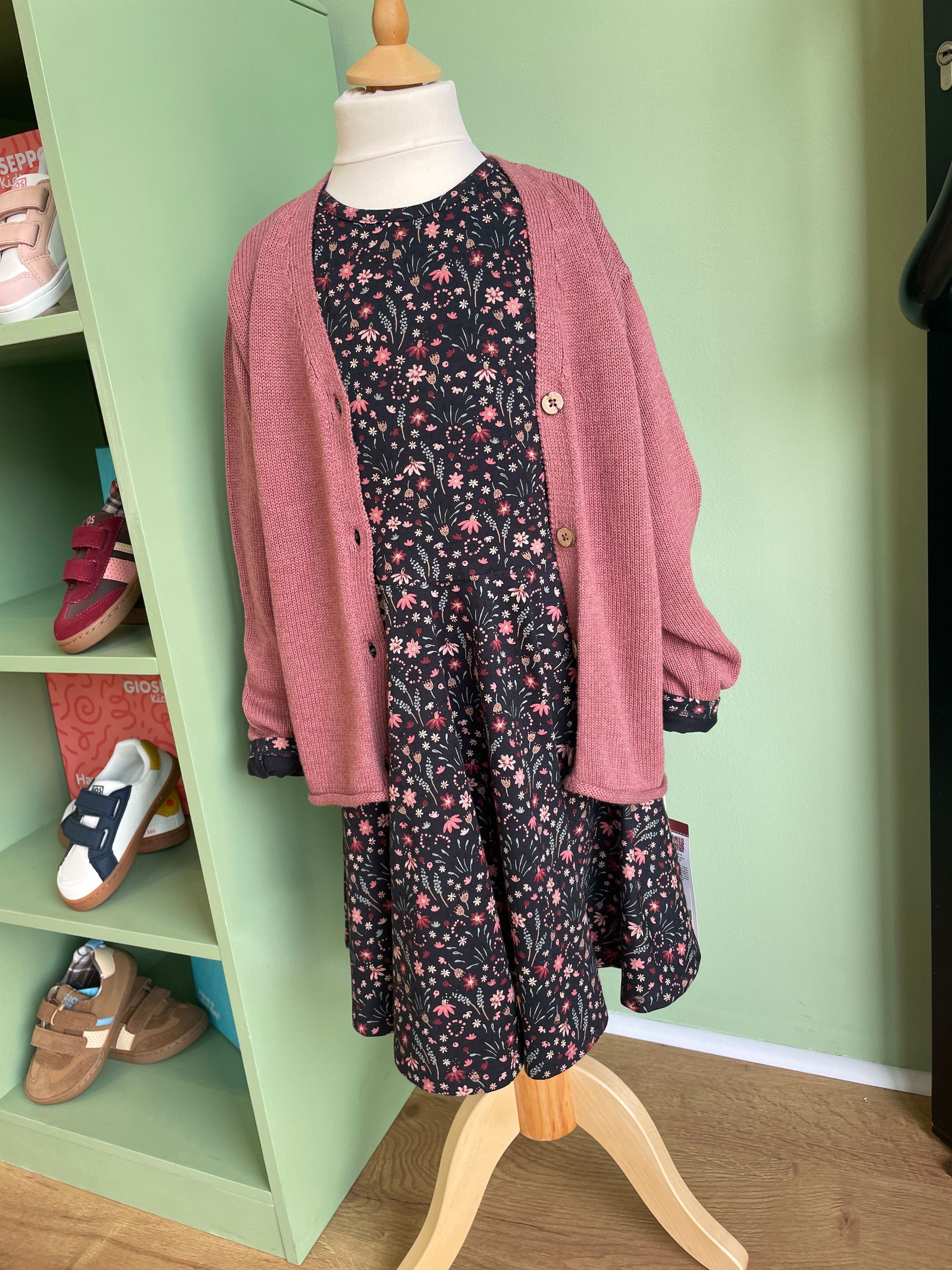 Abito a fiori con cardigan rosa su un manichino in una stanza verde, esposto su un supporto di legno