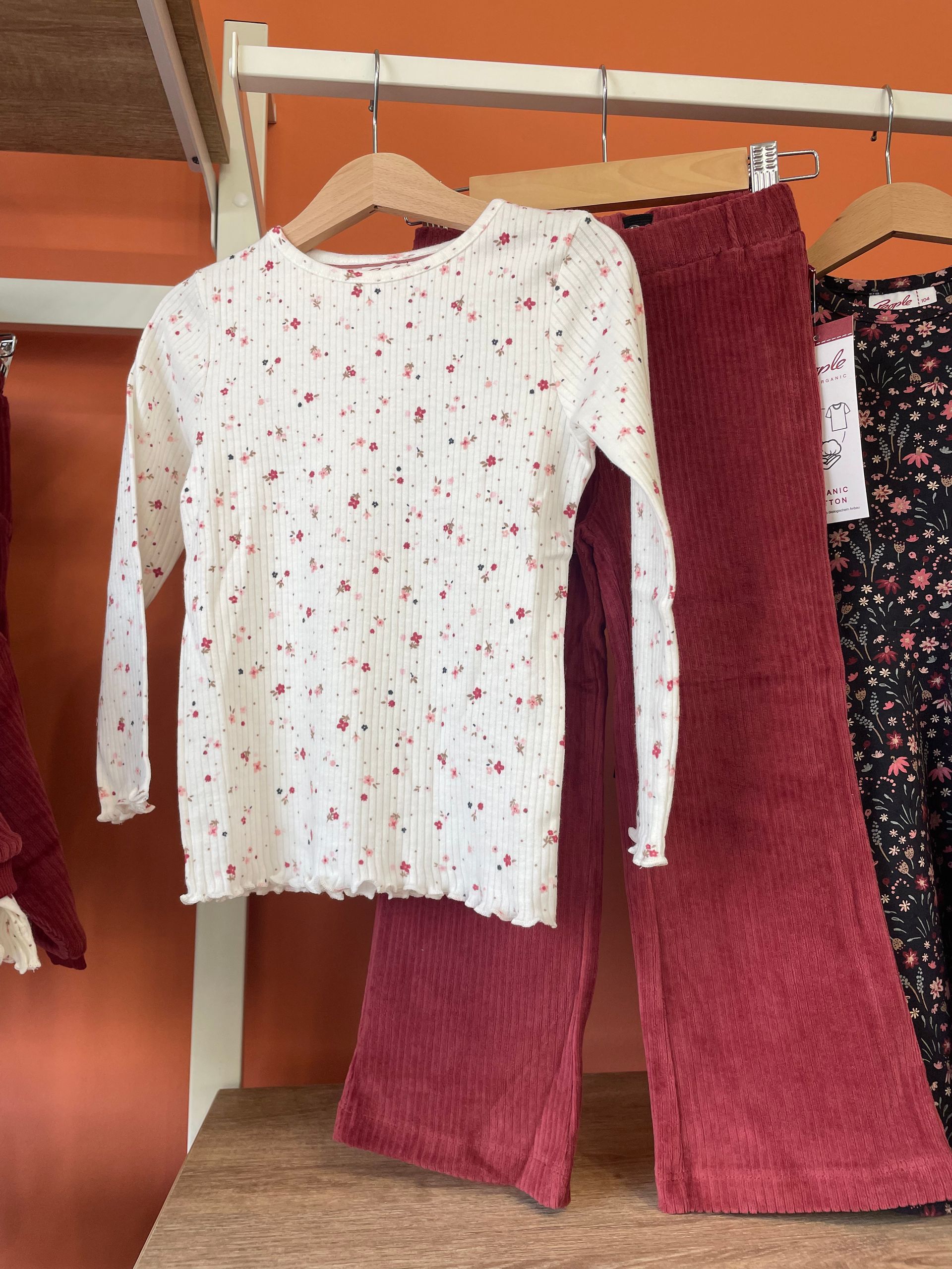 Maglia bianca a maniche lunghe con una piccola stampa rossa appesa accanto a pantaloni bordeaux su un appendiabiti
