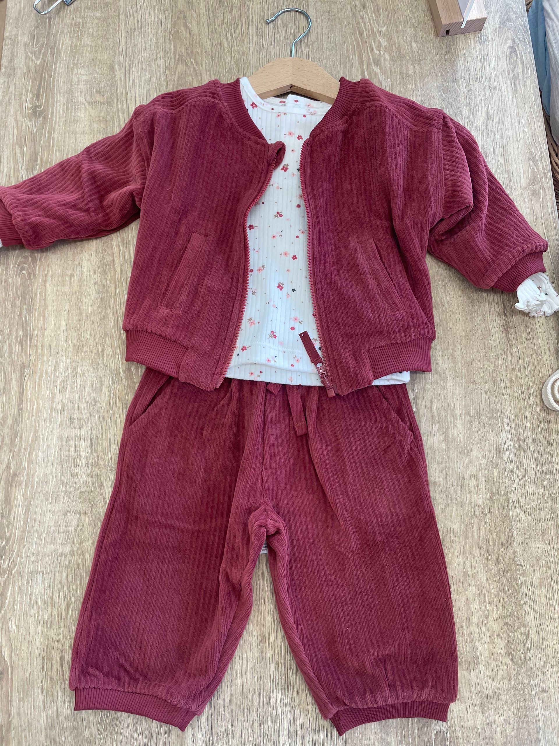 Completo composto da giacca e pantaloni bordeaux in morbido tessuto peloso, adagiato su una superficie beige texturizzata, con una camicia bianca a puntini sotto