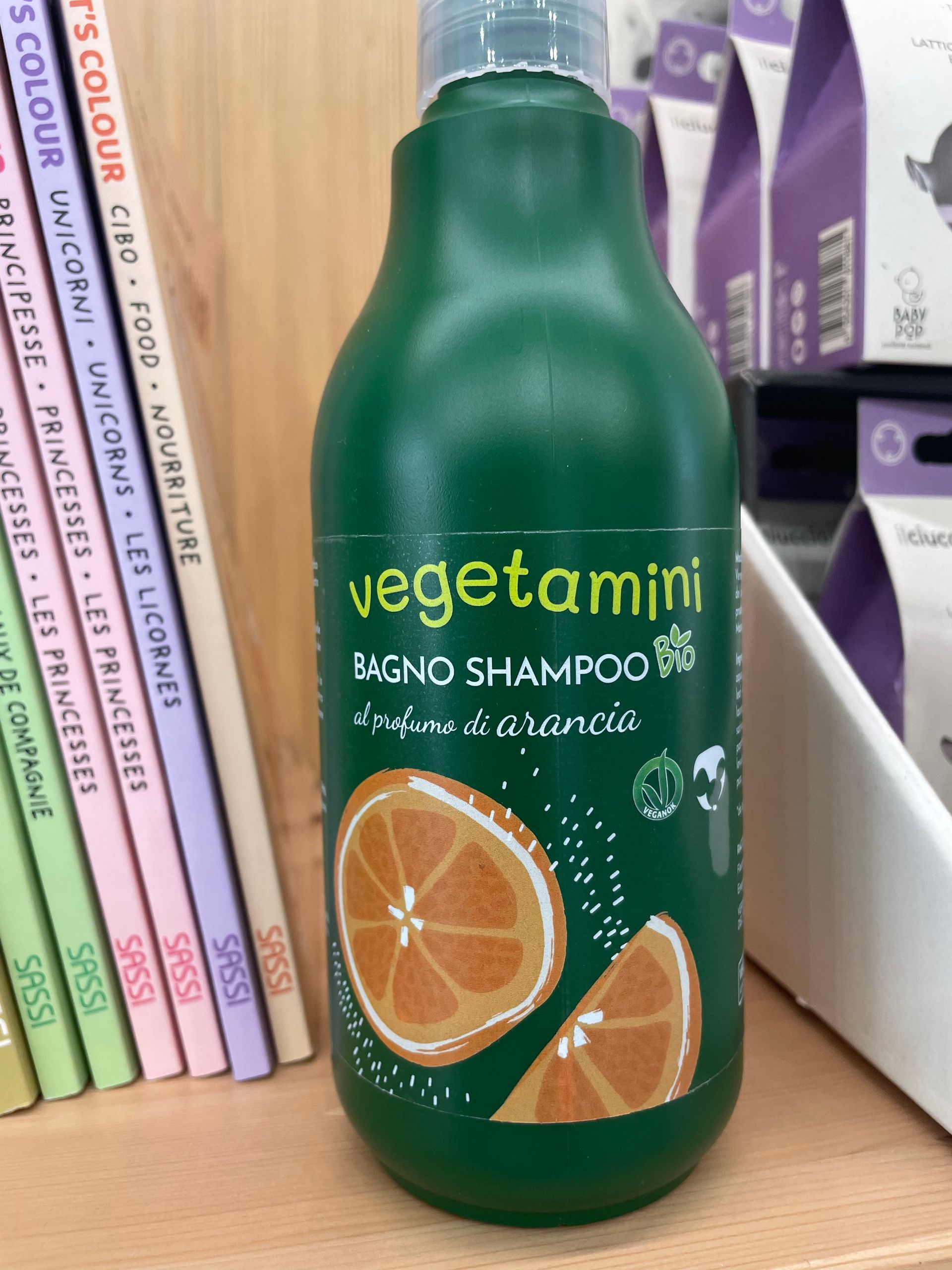 Flacone di shampoo Green Vegetamin con fette d’arancia su una mensola accanto ai libri.