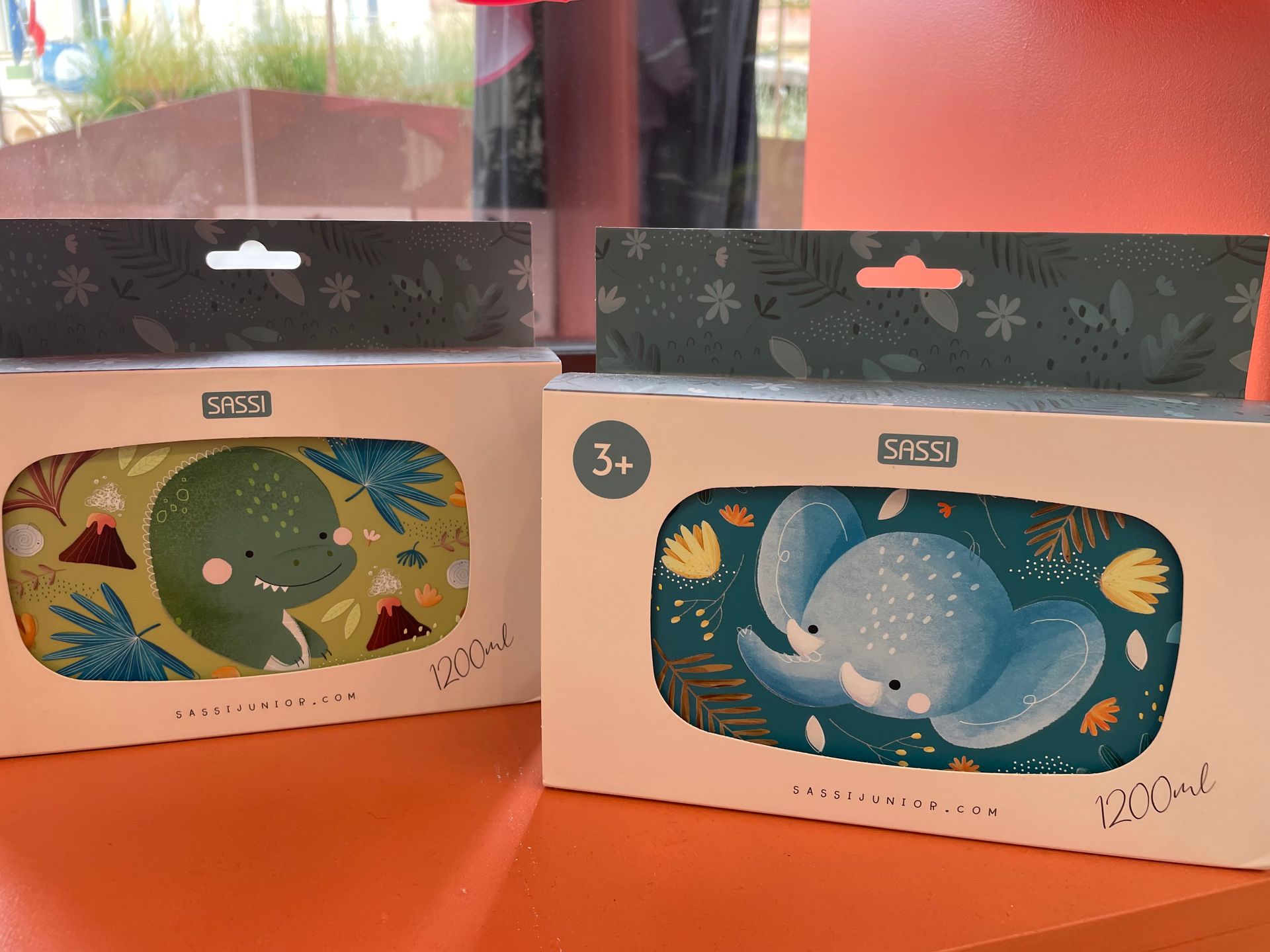 Due prodotti per bambini colorati e confezionati in scatole, con decorazioni a foglie e farfalle, su un tavolo.
