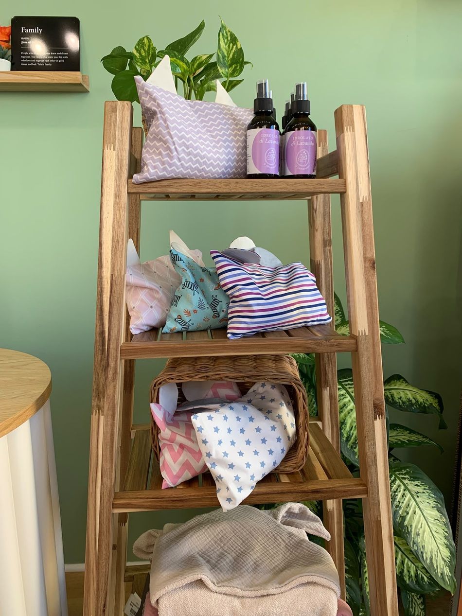 Scaffale in legno con coperte piegate, cuscini e flaconi di lavanda davanti a una parete verde con piante.