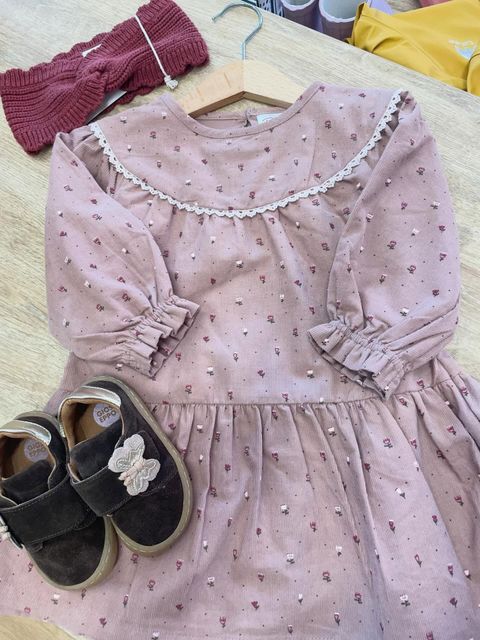 Vestito rosa da bambina con stampa floreale, abbinato a scarpe nere e a un capo in maglia bordeaux appeso a una gruccia.