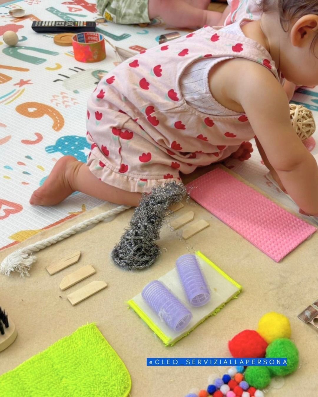 Bambino piccolo inginocchiato su un tappetino da gioco, mentre cerca di raggiungere giocattoli sensoriali colorati e oggetti dalle diverse consistenze.