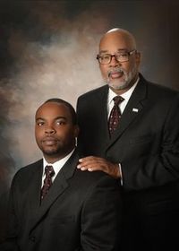 Two men in dark suits pose formally against a studio backdrop, one standing behind with a hand on the other's shoulder.