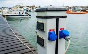 A dock with boats docked in the water and a pole with electrical outlets attached to it.