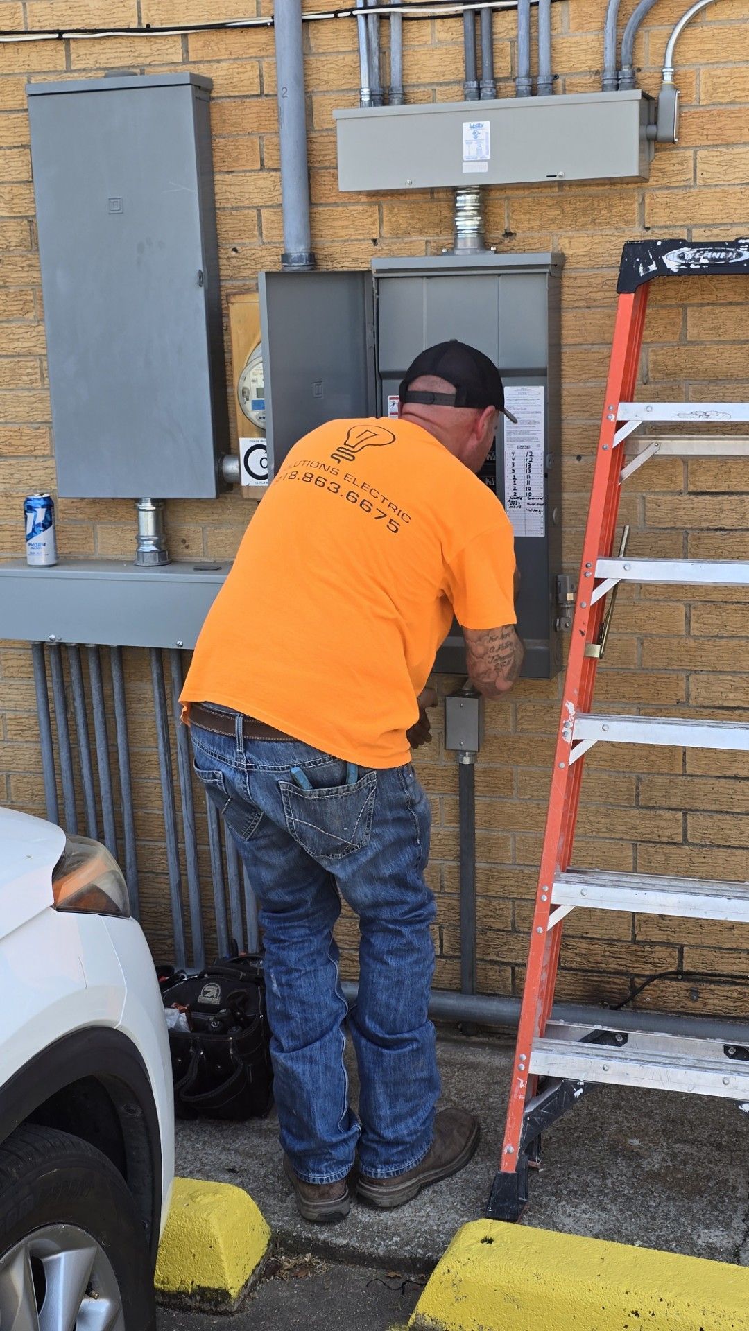 Worker in an orange shirt and jeans working on an electrical panel beside a ladder outdoors.