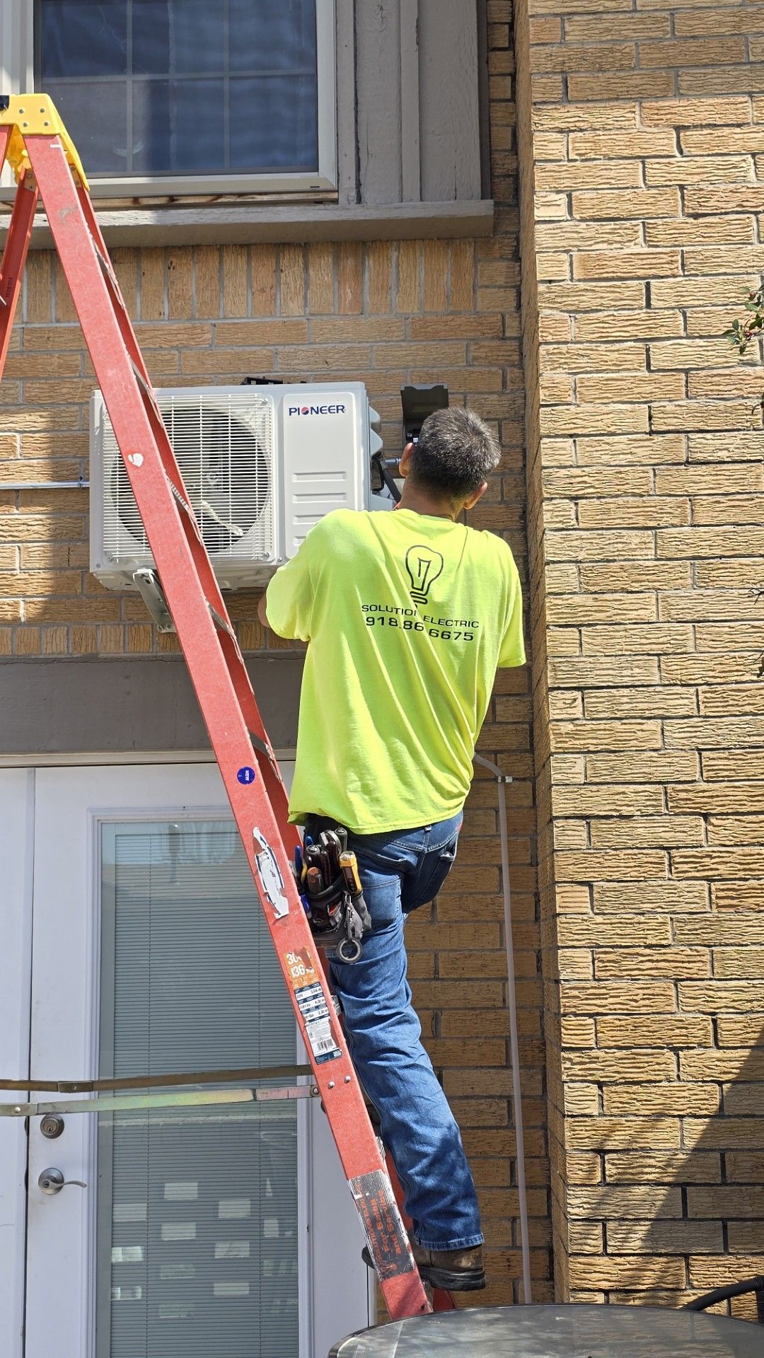 Worker on a red ladder installing an air conditioner on a brick building wall.