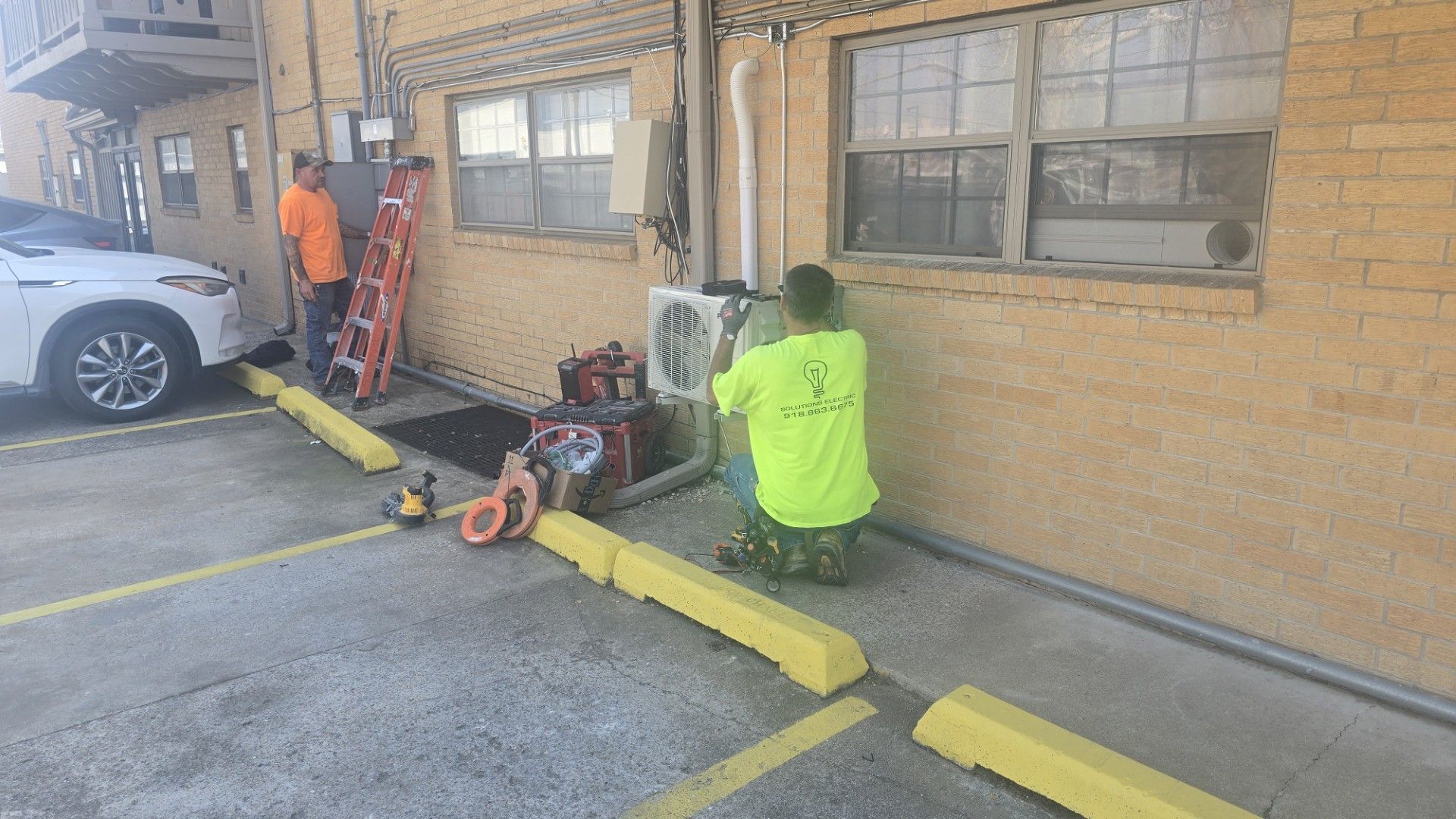 Workers servicing equipment beside a tan building; one kneels in a neon shirt while another stands by an open door.