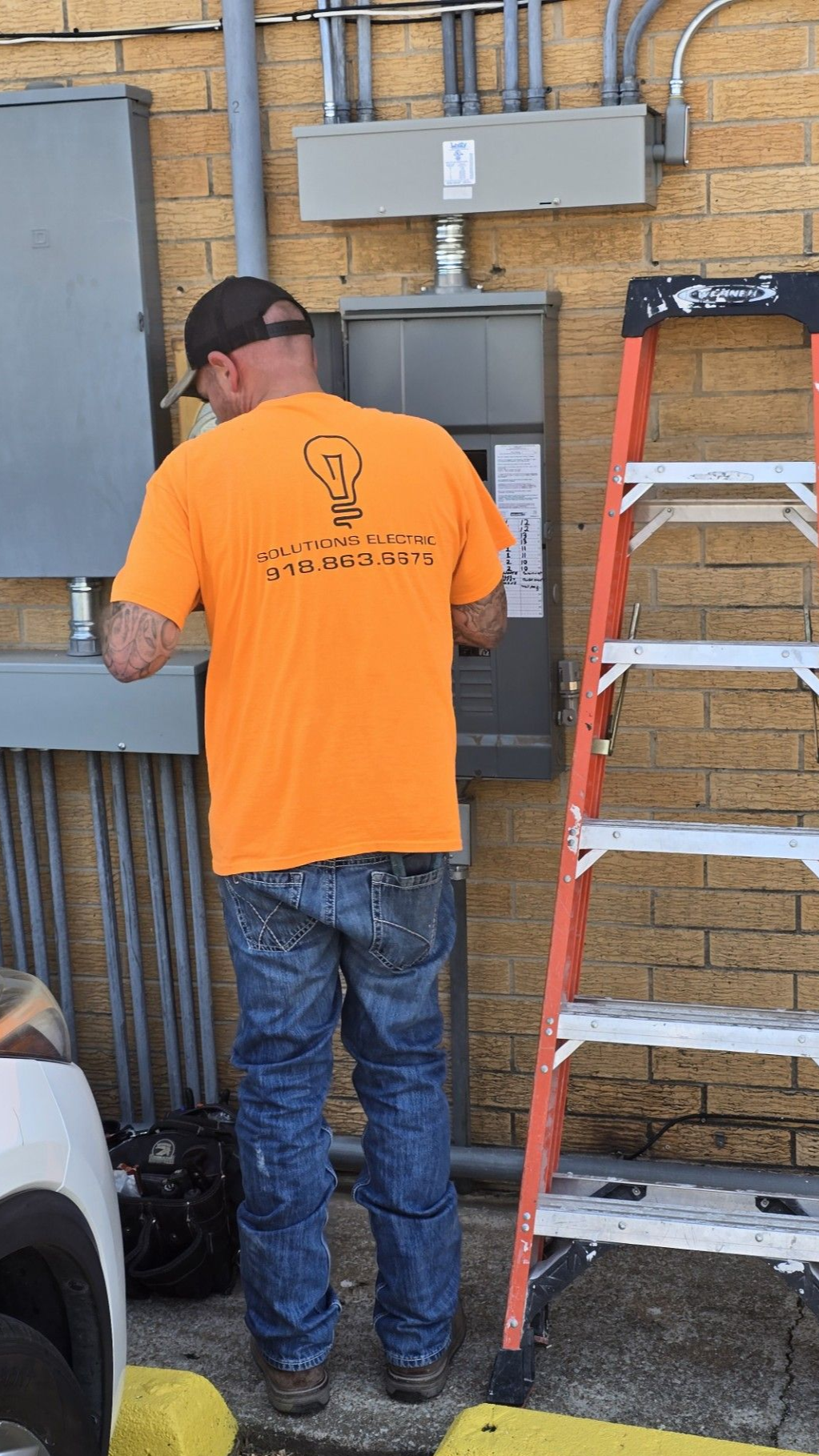 Worker in orange shirt stands at a railing beside a red ladder, facing a brick wall.