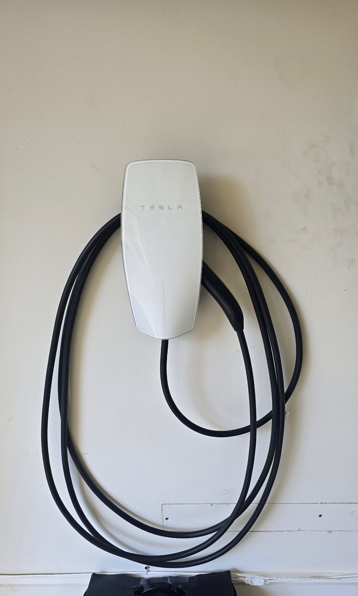 A white Tesla Wall Connector electric vehicle charger mounted on a wall with its black charging cable coiled around it.