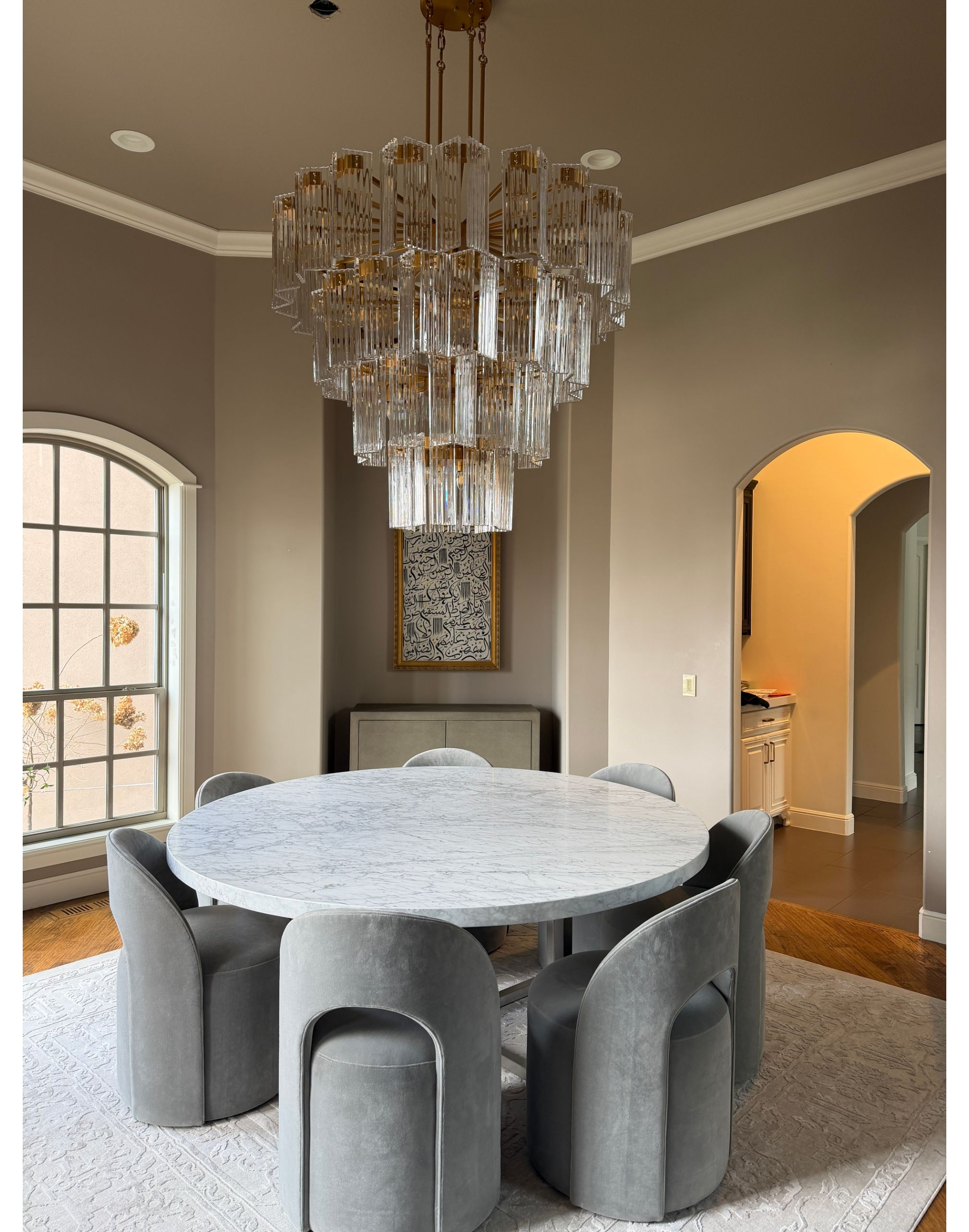 A round marble dining table surrounded by six grey chairs, topped with a tiered crystal chandelier in a neutral room.