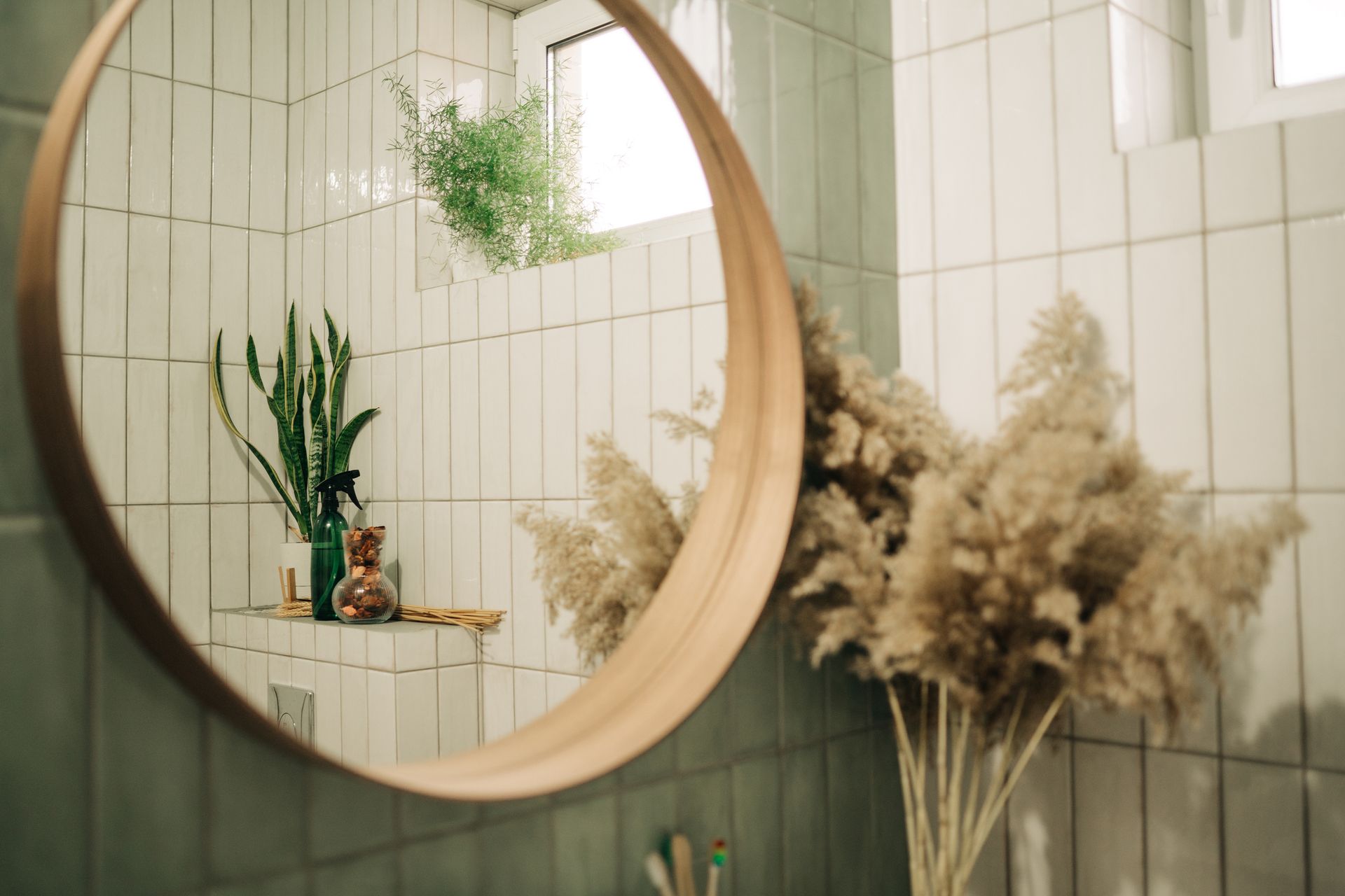 Round mirror reflecting plants and a shelf, with a vase of fluffy beige pampas grass in the foreground, set in a tiled bathroom.