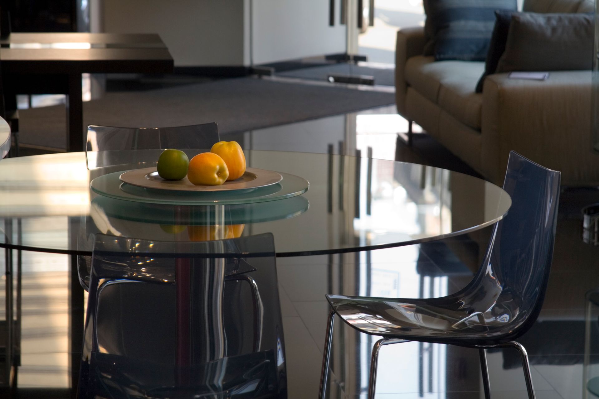 A modern dining room with a glass table, clear chair, and a bowl of fruit. Sunlight reflects off the glossy floor.