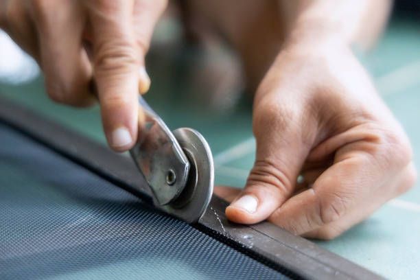 Person using a roller tool to secure screen mesh into a window frame's groove. Close-up shows hands, tool, black mesh, and a dark gray frame.