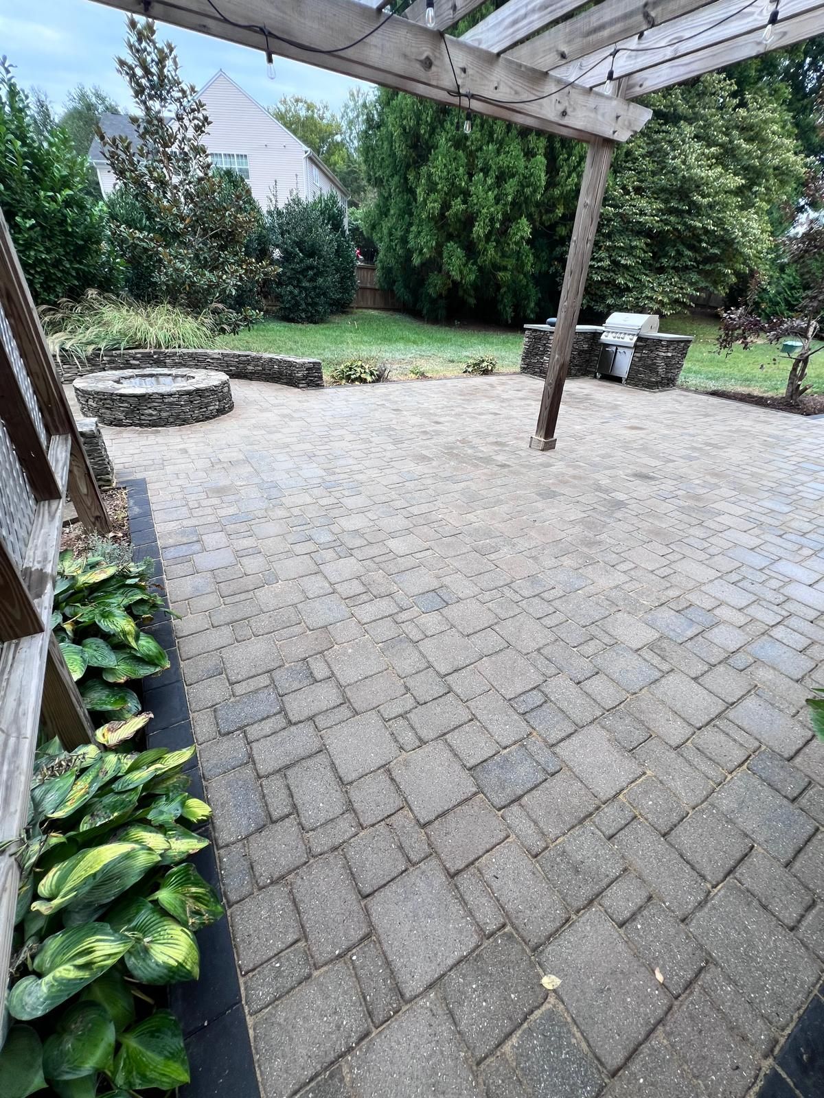 Brick patio with a fire pit, grill, and pergola; green plants in the foreground.