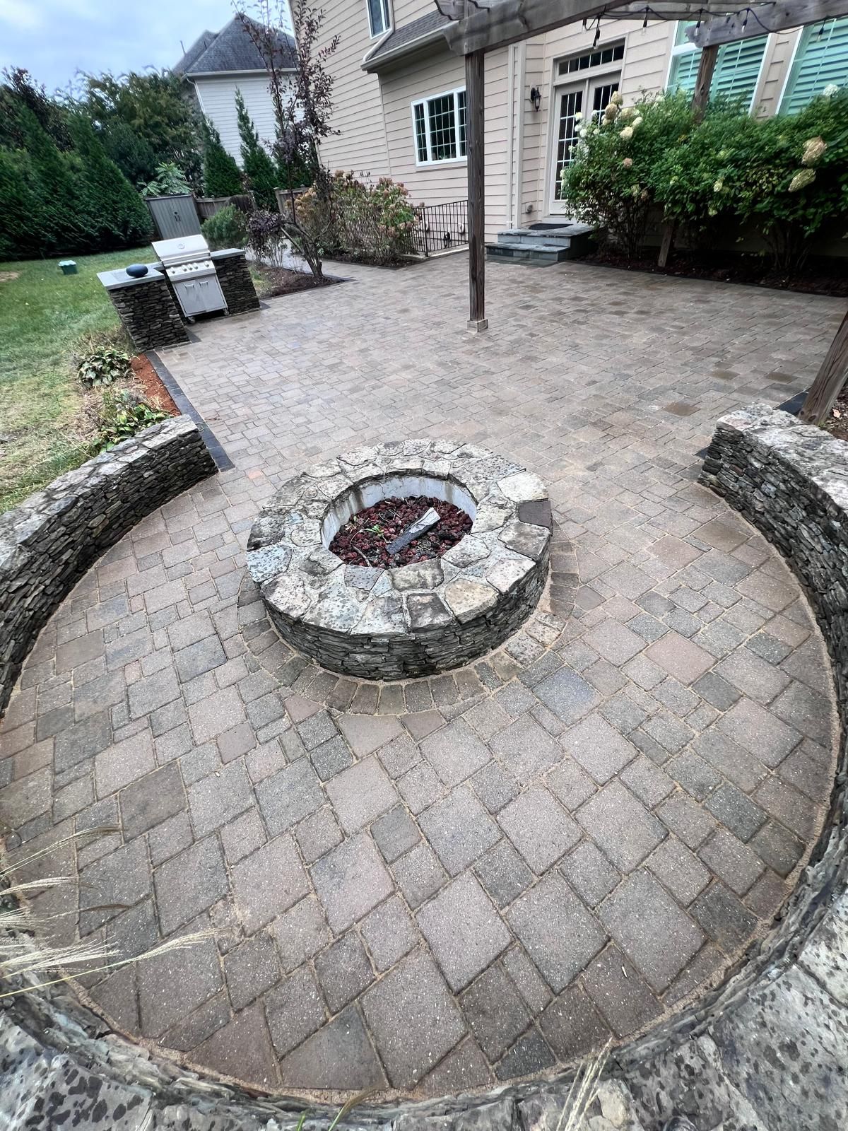 Circular stone patio with fire pit, seating wall, and grill in backyard.