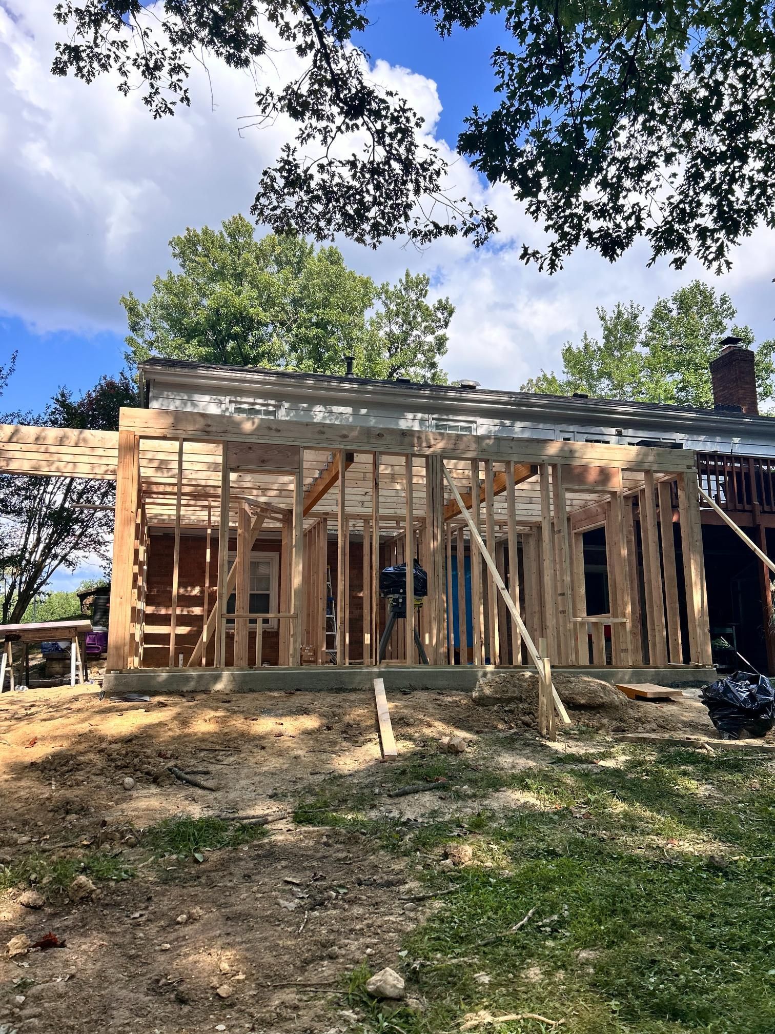 Wooden frame of a building under construction, attached to a house with green trees overhead on a sunny day.