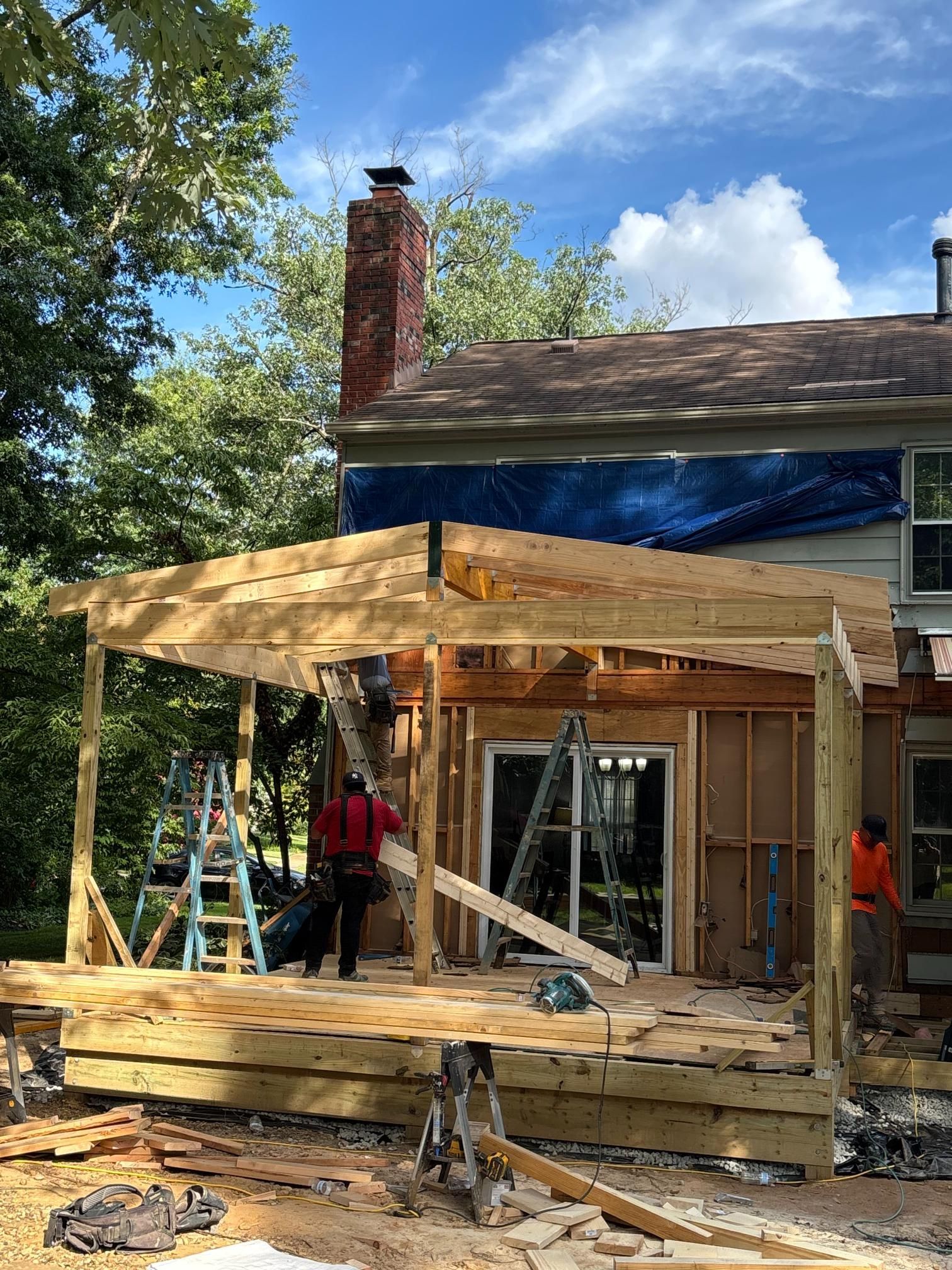 Construction of a wooden deck and roof addition to a house. Workers are on the deck, with tools, ladders.