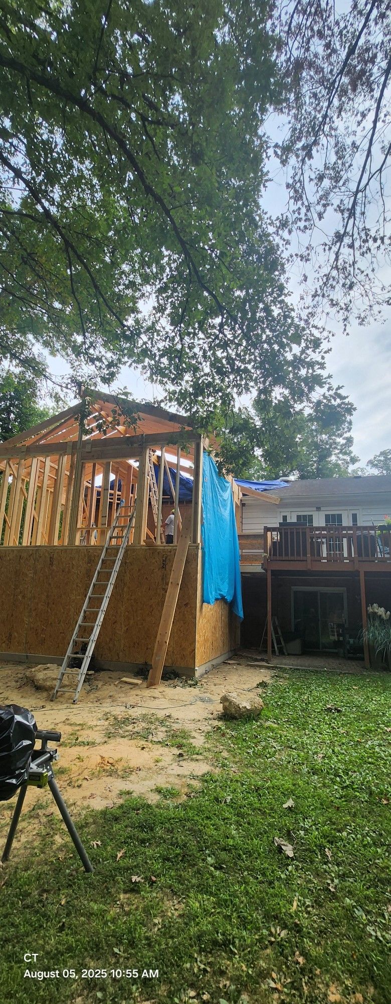 A wooden structure under construction with a ladder, covered in a blue tarp, and a wooden deck in the background.