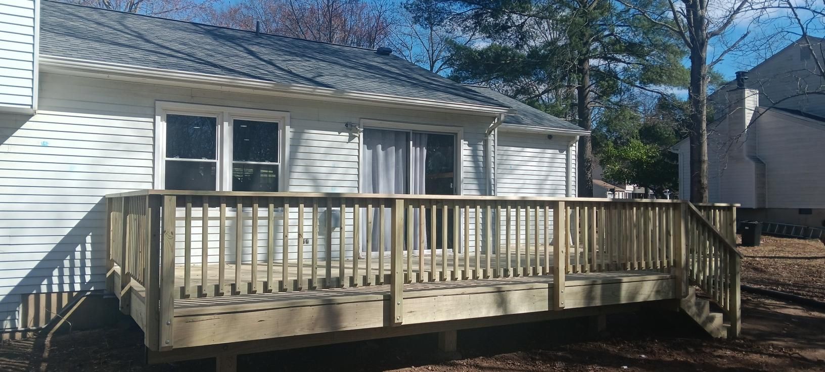 Wooden deck outside a white house with a sliding door and trees. Blue sky.