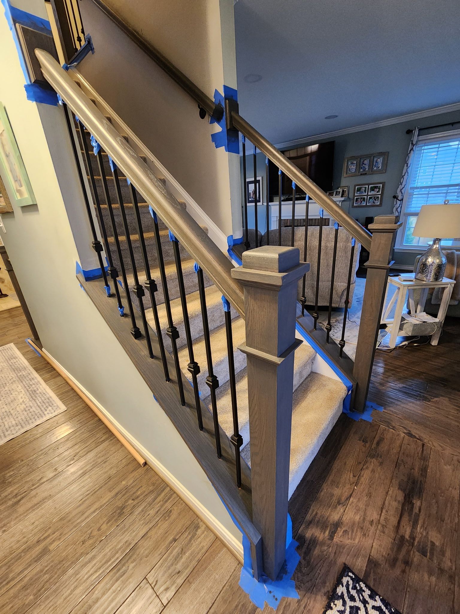 Staircase with gray wooden handrails and black metal spindles. Blue tape protects adjacent surfaces.