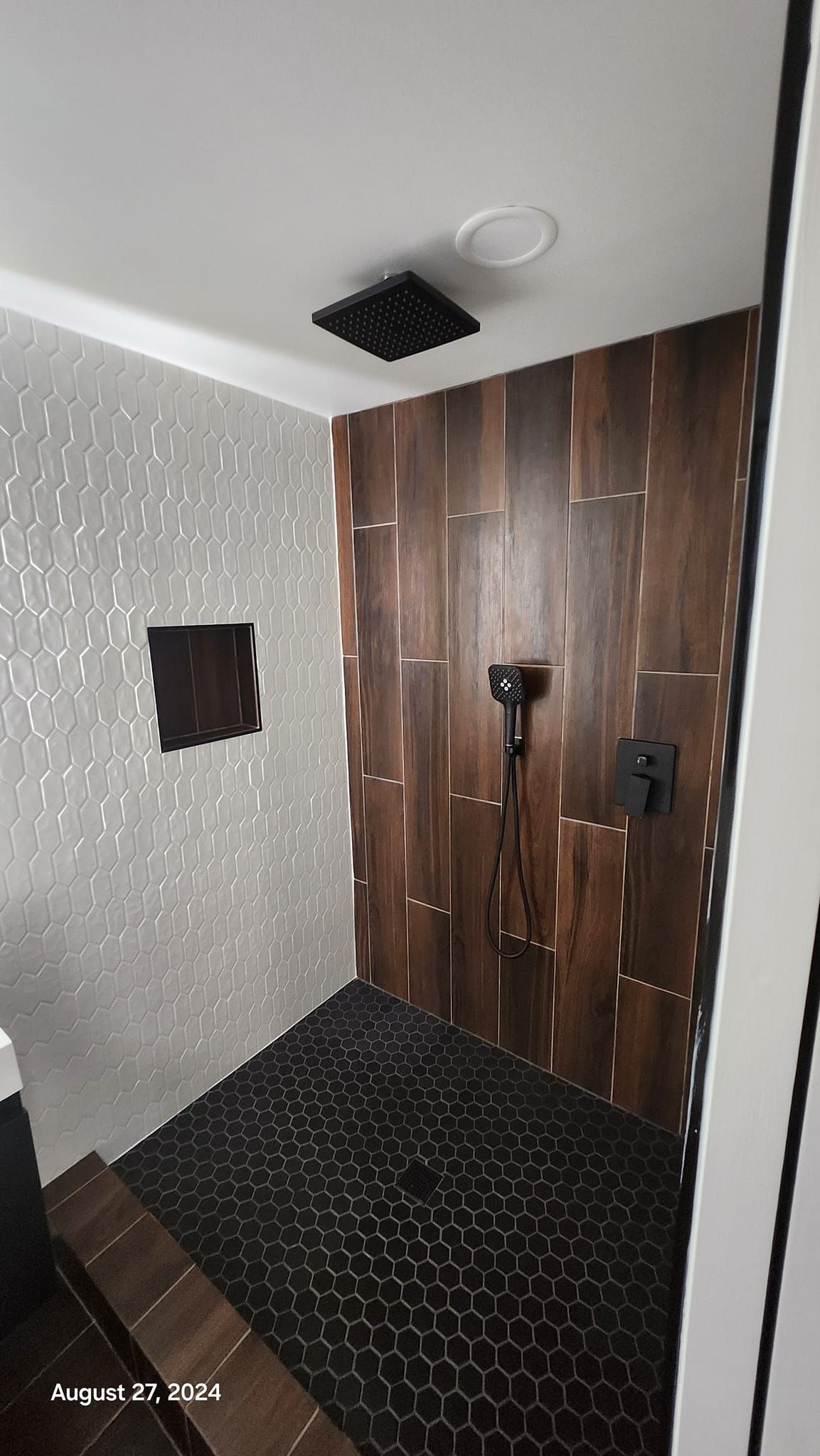 Shower with wood-look and white tile walls, black pebble floor, black shower fixtures, and a black square shower head.