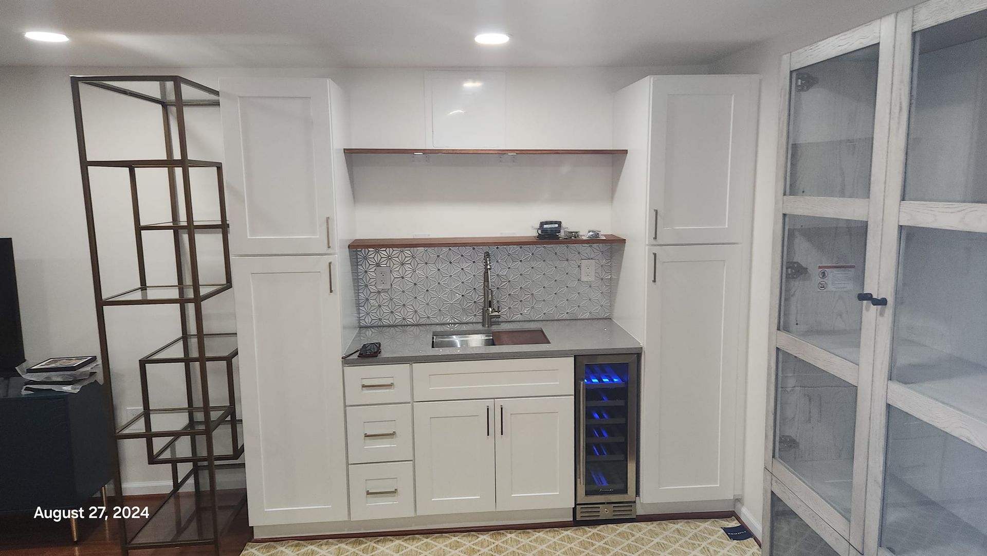 Small white bar area with sink, wine cooler, cabinets, mosaic backsplash, and metal shelving.