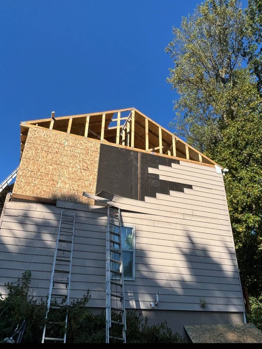House under construction; roof framing, siding removal, ladders against the wall, clear sky, and foliage.