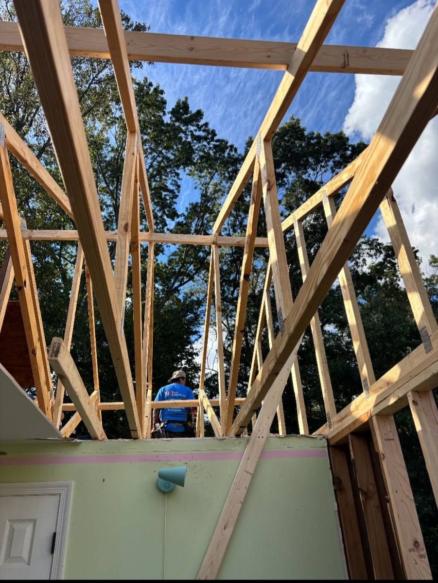 Wood framing of a roof under construction, with a worker visible; blue sky and trees in background.
