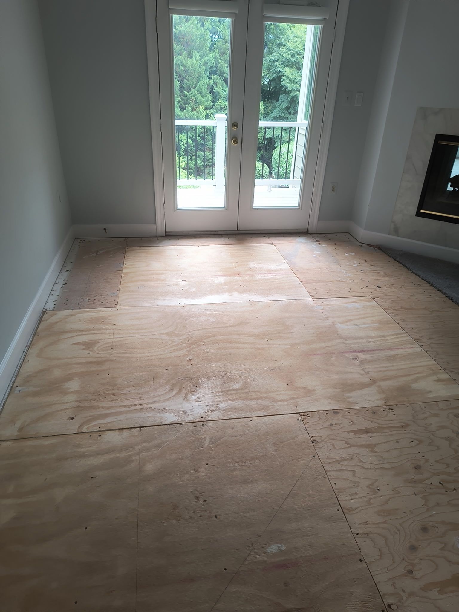 Empty room with plywood subfloor and French doors opening to a bright patio.