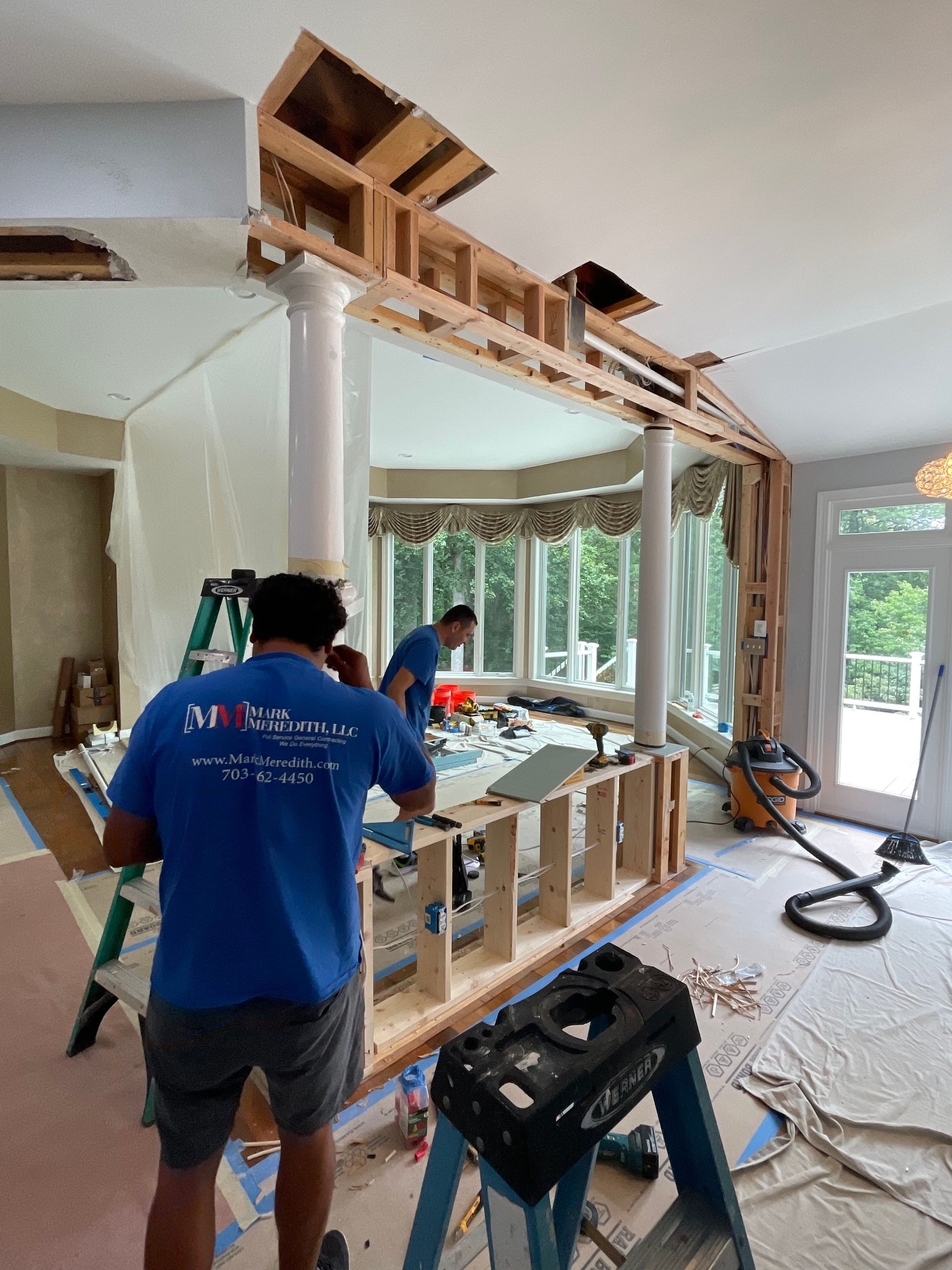 Two workers framing an indoor wall with exposed wood studs in a bright, unfinished room.