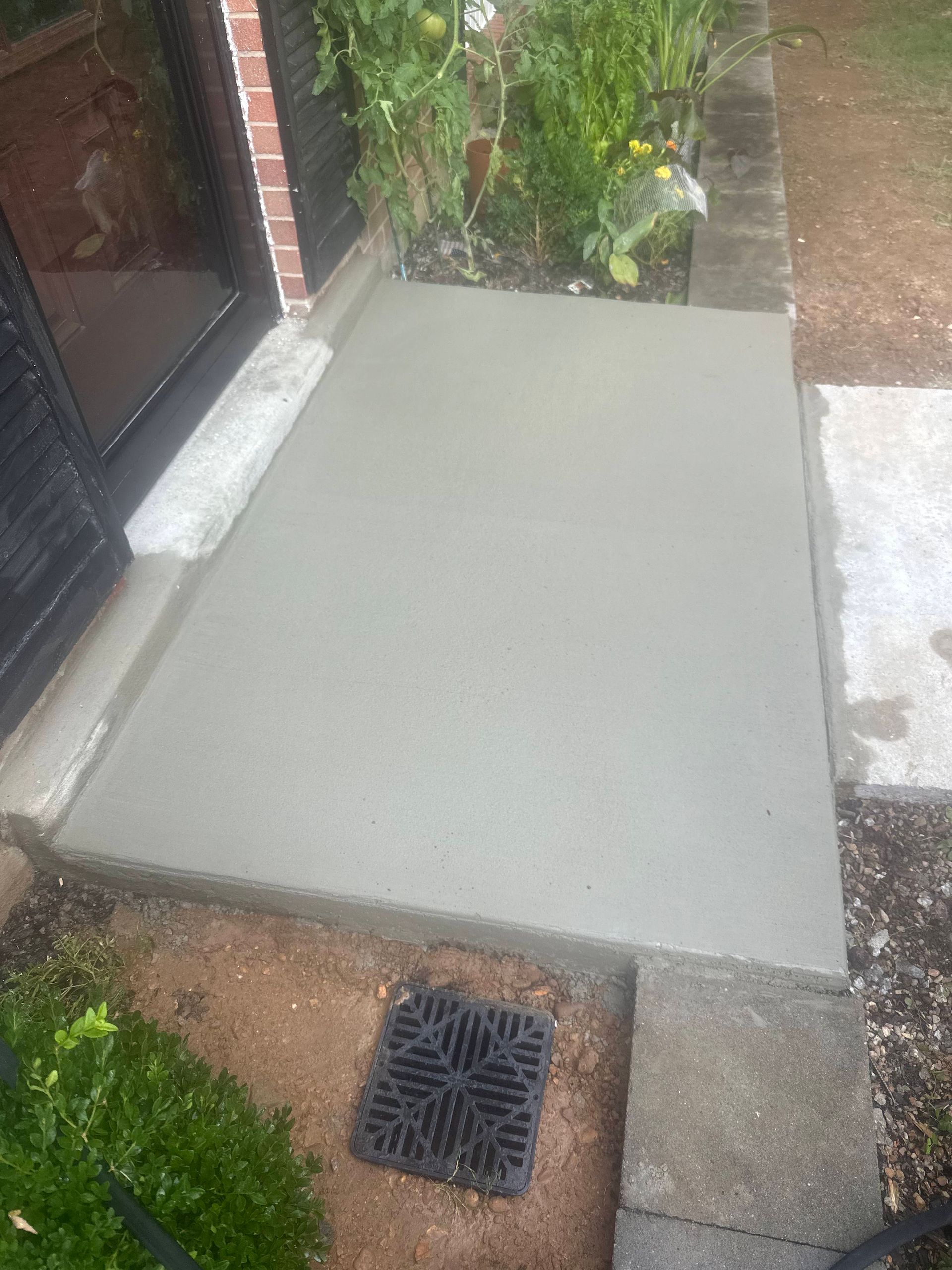 Freshly poured concrete patio slab beside a brick house with a drain grate and garden border.