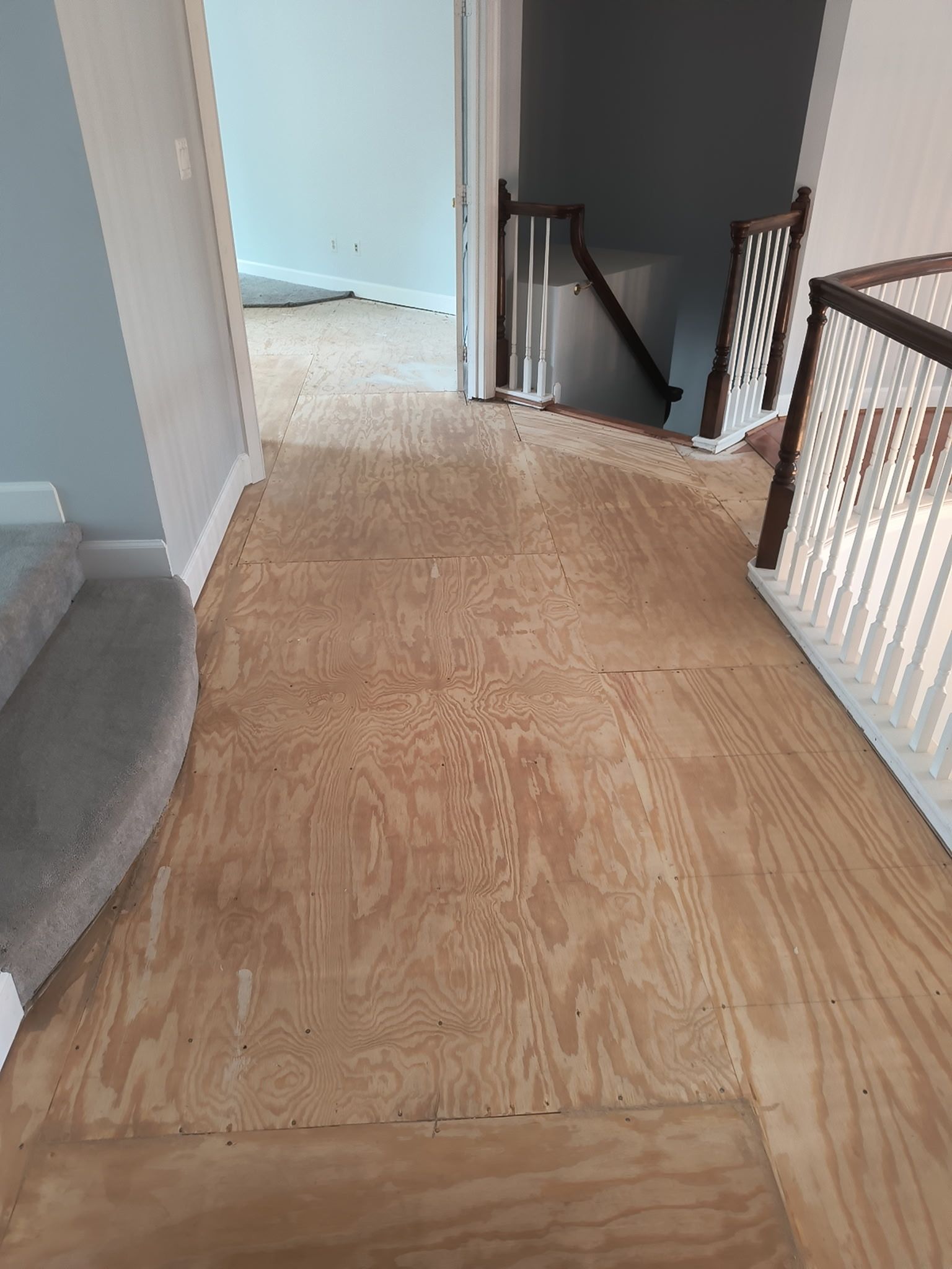 Hallway with plywood subfloor, gray carpet edge, and staircase railing leading to a bright room