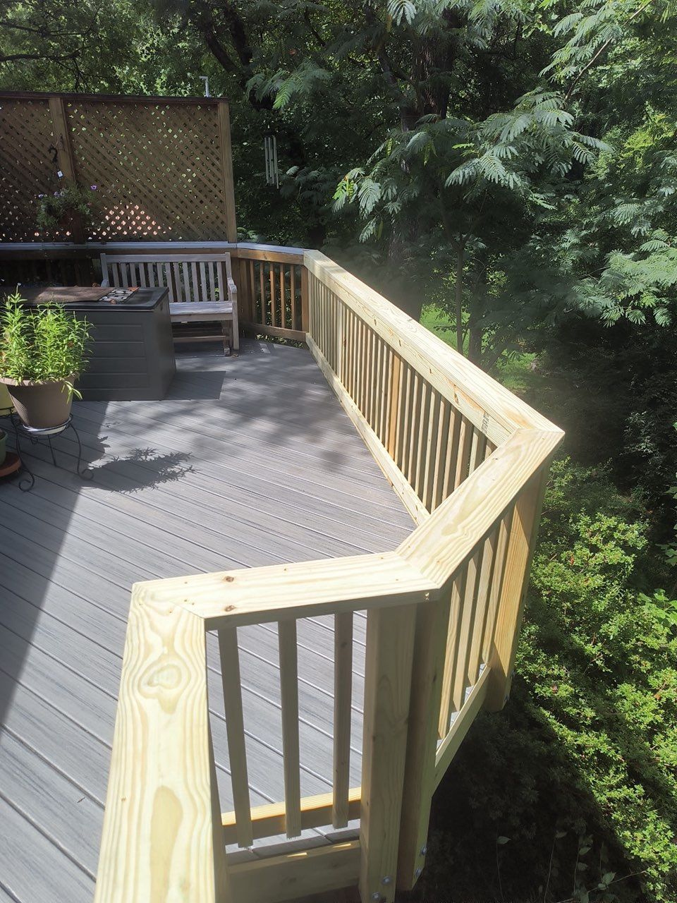Wooden deck with railing, potted plants, and outdoor seating beside dense greenery.