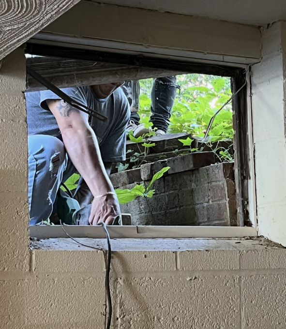 Person working at a basement window. They are reaching into the window, with foliage visible outside.